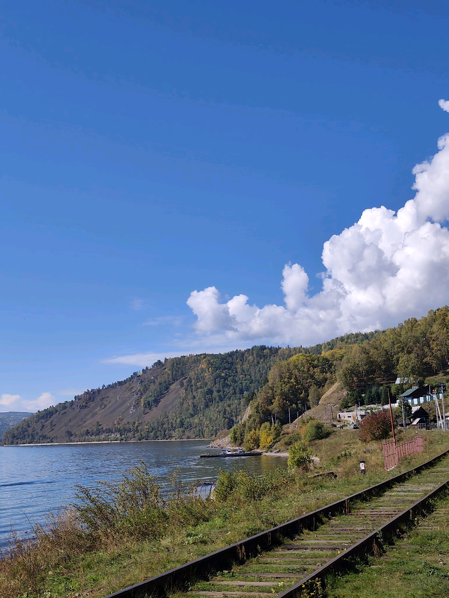 The image depicts a picturesque view of Lake Baikal in Russia, captured on a clear, sunny day. In the foreground, a set of disused train tracks runs diagonally across the frame, overgrown with green grass and some yellow wildflowers. To the left, the calm, blue waters of Lake Baikal stretch towards a distant, tree-covered mountain range. The slopes of the mountain are a mix of green evergreen trees and autumnal deciduous trees displaying yellow and orange foliage.
Along the shoreline, a few small buildings are visible nestled amongst the trees, suggesting a small settlement or a remote outpost. There are also a couple of boats moored at a small dock. The sky is a vibrant blue, dotted with fluffy white cumulus clouds, particularly prominent on the right side of the image. The overall scene conveys a sense of tranquility and natural beauty, with the juxtaposition of man-made infrastructure (the train tracks) against a wild, natural landscape. No people are clearly visible in the image.