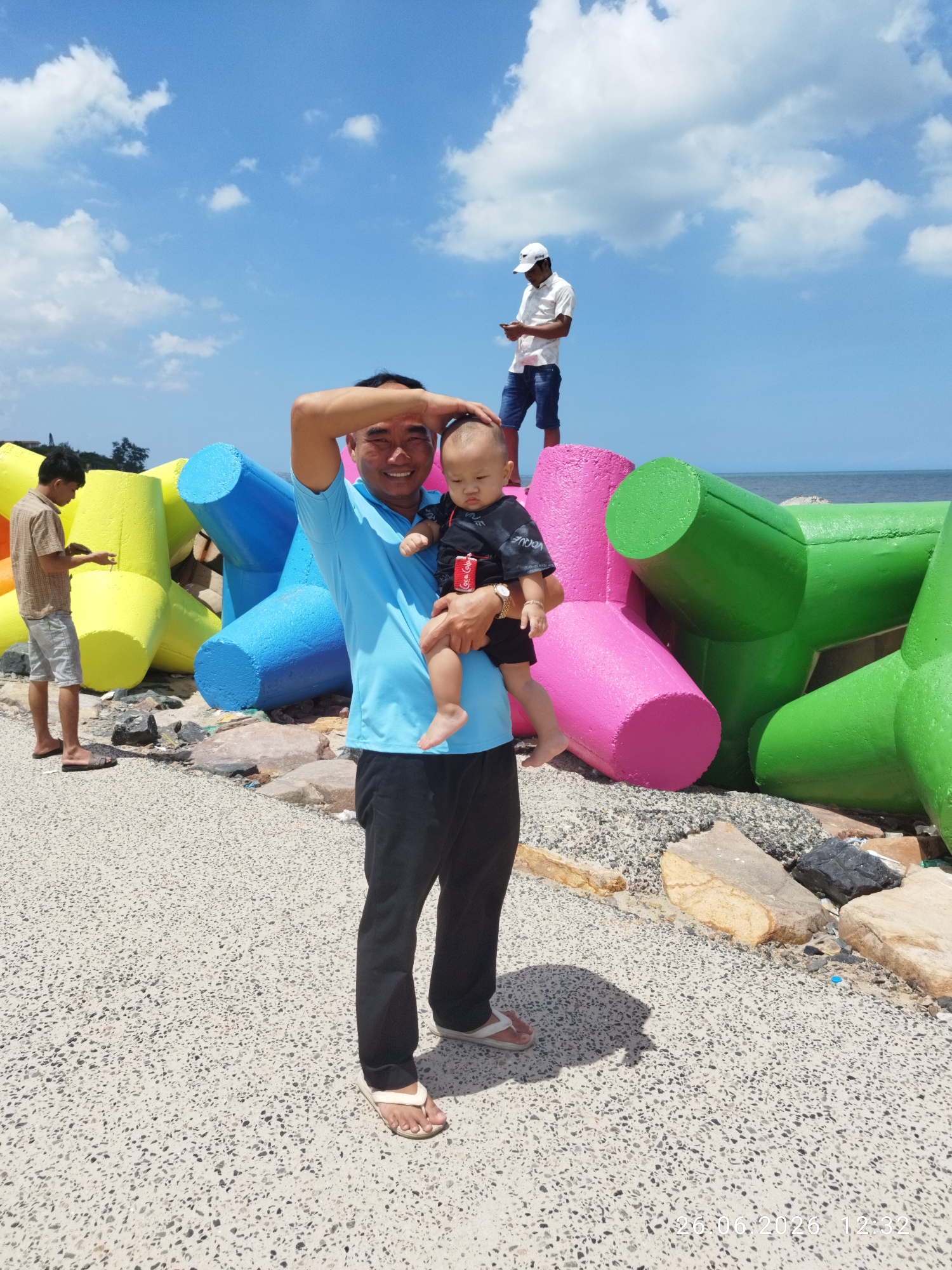 The image captures a sunny day at a beach, likely in Phu Thuy Ward, Vietnam, given the contextual information. In the foreground, a man in a blue t-shirt and black pants holds a baby. He is smiling and has his hand on the baby's head. The baby is wearing a black t-shirt. To the left of the main subjects, another man in a patterned shirt and shorts is looking at his phone. In the background, a third person, wearing a white shirt and a white cap, stands on a colorful structure made of large concrete tetrapods painted in bright yellow, blue, pink, and green. These tetrapods appear to form a breakwater or artificial reef along the coastline, where the sea is visible under a bright blue sky with scattered white clouds. The ground in the foreground is a textured, light-colored surface, possibly a pathway or boardwalk. The overall mood is relaxed and cheerful, indicative of a casual day out by the sea. A timestamp at the bottom right of the image reads "26.06.2026 12:32".
