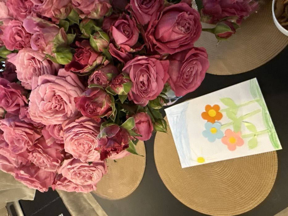 A high-angle shot displays a bouquet of pink roses, some in full bloom and others as buds, situated on a dark surface with round, woven placemats. A child's drawing of flowers with colorful petals and green stems rests on one placemat. The backdrop is a dark, possibly wooden, surface with a bowl visible in the upper right. The composition focuses on the roses and the artwork, creating a warm, domestic scene.