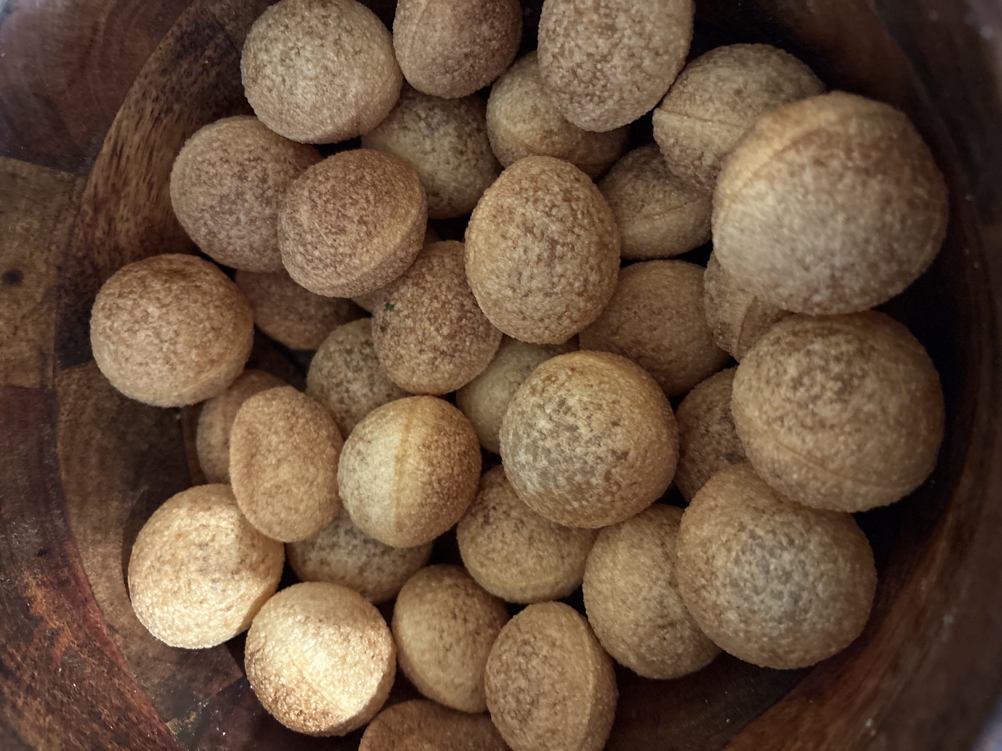 A close-up, top-down shot showcases a dark brown, wooden bowl filled with numerous small, round, golden-brown savory snacks. The snacks have a slightly textured surface and appear to be uniform in size, with some showing a subtle line around their circumference. The bowl's inner surface is visible along the edges of the frame, its wood grain creating a rustic backdrop. The lighting highlights the convex surfaces of the snacks, casting soft shadows and giving them a slightly luminous quality. The image is static, with no movement or action depicted. The setting is indoors, as indicated by the enclosed nature of the bowl and the absence of any outdoor elements. There are no people visible in the image.