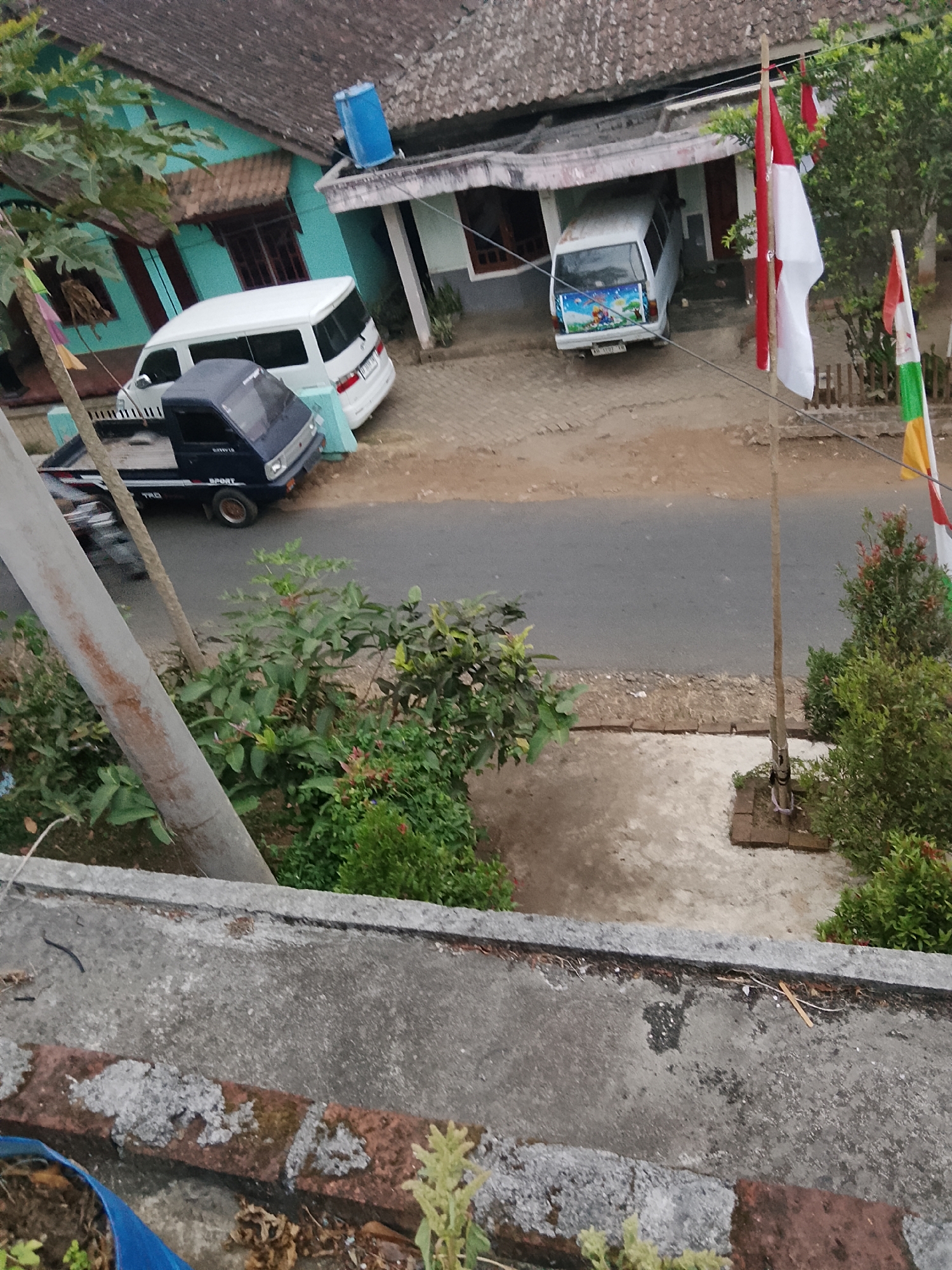 The image shows a street scene in Betonan, Indonesia. In the foreground, a grey concrete ledge is visible, with some loose debris and a blue plastic bag. Beyond the ledge, a paved road runs horizontally across the image. On the left side of the road, there is a dark blue pickup truck parked. Further down the road, a white van is parked in front of a building with a blue water tank on its roof. Another white van, decorated with cartoon characters on its rear, is parked in a covered area to the right. Several flags are visible: a red and white Indonesian flag on a pole on the right, and other flags with red, white, and green sections. Lush green vegetation is present on both sides of the road. The scene appears to be a residential area with modest buildings.