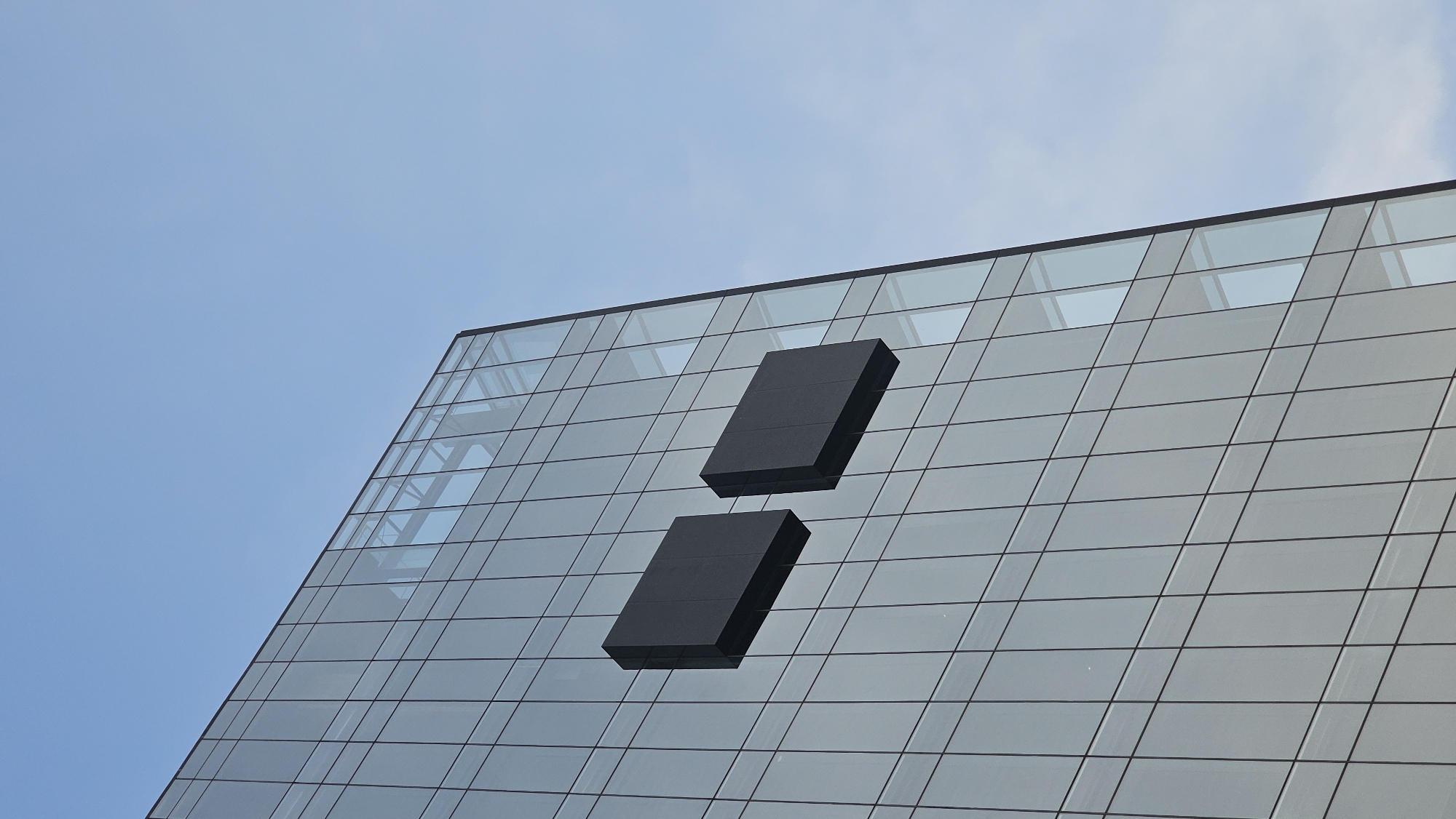 The image shows the upper portion of a modern building in Puebla, Mexico, constructed primarily of glass and metal. The glass panels are arranged in a grid-like pattern, reflecting the blue sky. Two dark, rectangular elements protrude from the facade, positioned vertically with a space between them, forming a design element. The sky is visible in the background, appearing light blue with a few faint clouds. The angle of the photograph suggests it was taken from below, looking up at the building.