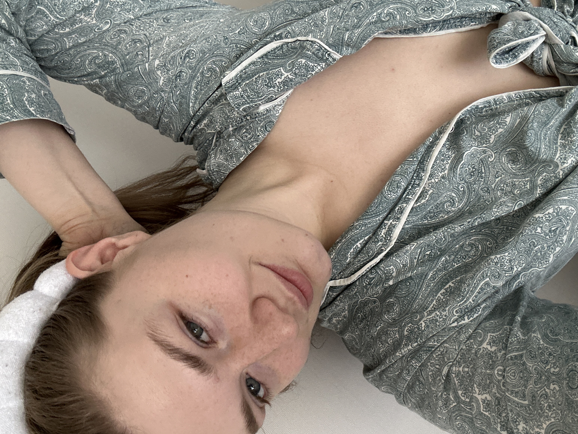 A young woman with fair skin, freckles, and brown hair is lying down, possibly on a white surface. She is wearing a blue and white paisley-patterned pajama top with white piping. She has a white headband. Her gaze is directed towards the camera. The location is not identifiable as Kazan, Russia based on the provided image.