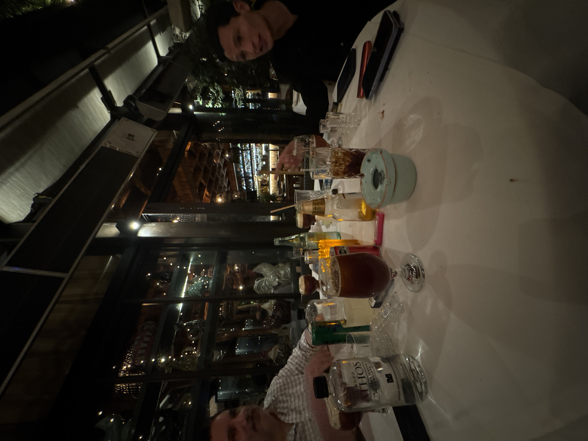 The image shows a scene at a bar or restaurant in Mexico City, Mexico, at night. Two people are present. One person is wearing a black shirt and seated at the table. Another person is partially visible, wearing a white and gray patterned shirt, and also appears to be seated at the table. The table is covered with a white tablecloth and has numerous drinks and beverages in glasses and bottles on it. There is also an ashtray on the table. In the background, there is an illuminated structure with a glass window revealing decorative interior decorations behind the seated individuals.
