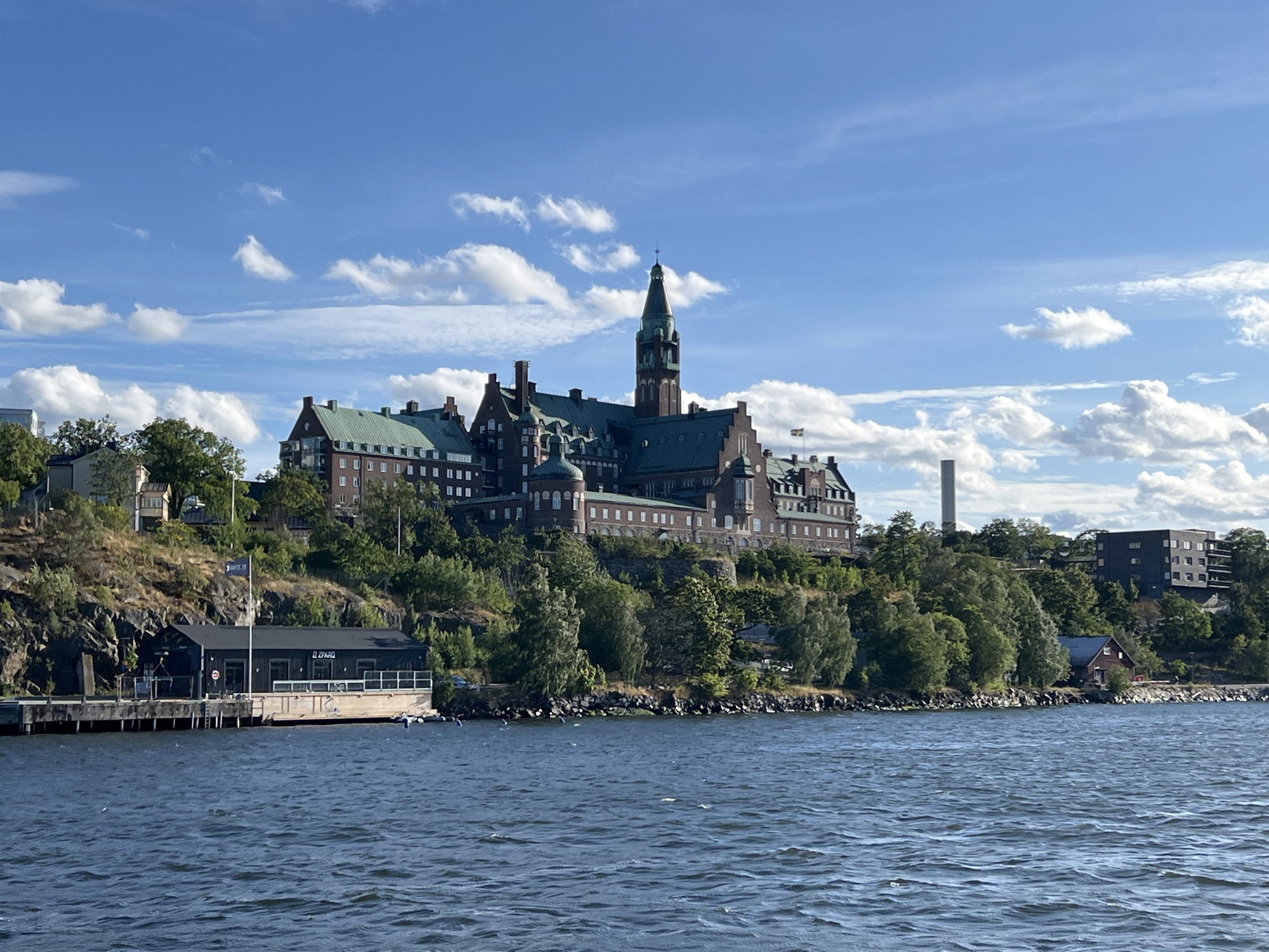 The image shows a large, historic building with a green roof and a prominent tower, situated on a hill overlooking a body of water. The building appears to be made of red brick and has many windows. It is surrounded by lush green trees. In the foreground, the water is choppy with visible ripples. On the left side of the image, there is a dark-colored building with a pier extending into the water. The sky is blue with scattered white clouds. A Swedish flag is visible flying from a flagpole on the hill. The location is identified as Nacka, Sweden.