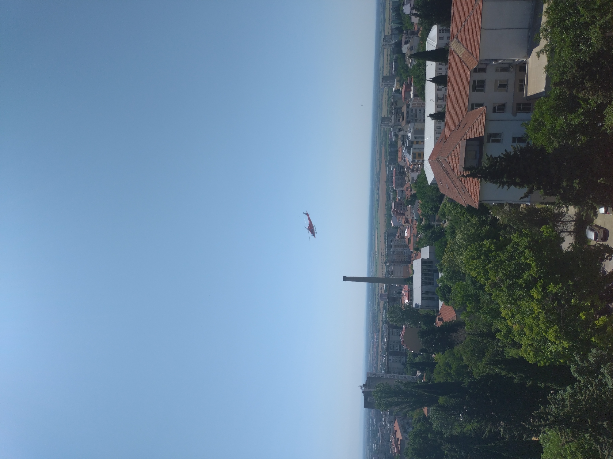 The image is an aerial view of the city of Stara Zagora, Bulgaria, on a clear, sunny day. In the sky, a red helicopter is visible, flying from left to right. Below, a cityscape unfolds with a mix of residential buildings, some taller apartment blocks, and industrial structures, including a prominent tall chimney. Green trees are abundant, interspersed between the buildings and in the foreground. In the lower right corner, a white building with a red tiled roof, possibly a hospital or an institutional building, is clearly visible. The background shows a flat horizon line, suggesting the city is situated in a plain. There are no visible people or specific interactions taking place.