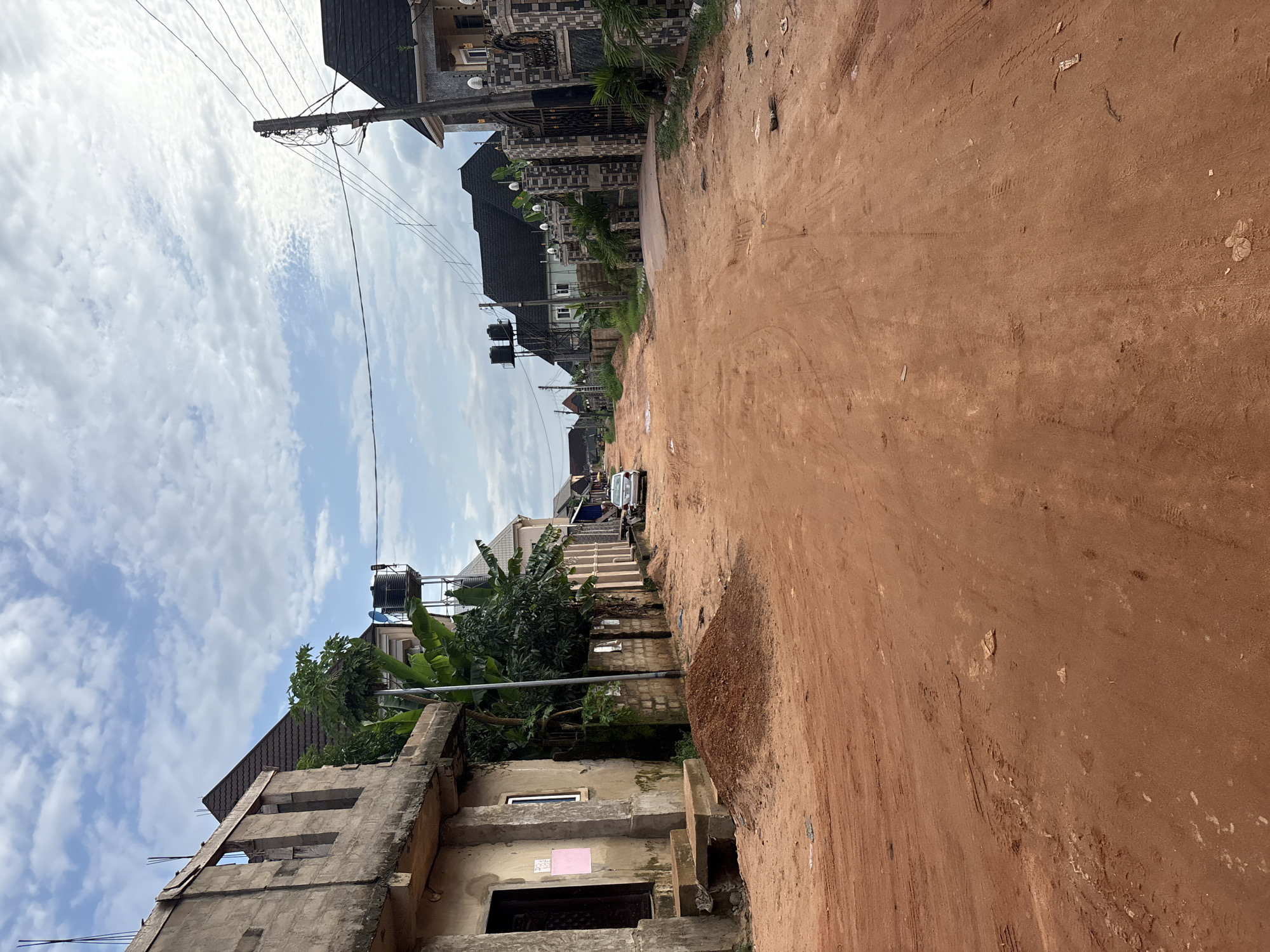 The image depicts a street scene in Obosi, Nigeria, characterized by a red dirt road under a cloudy sky. The road is uneven with tire tracks and patches of debris. On the left side of the road, there is an unfinished concrete building with exposed rebar and a dark doorway. Adjacent to it, a pile of red dirt sits on the roadside. Further down the left, more houses and lush green vegetation, including banana trees, are visible, along with elevated water tanks. On the right side of the road, houses with dark tiled roofs and decorative fences line the street. A silver sedan is parked on the right side of the road further down. The environment suggests a developing area with ongoing construction and unpaved infrastructure.