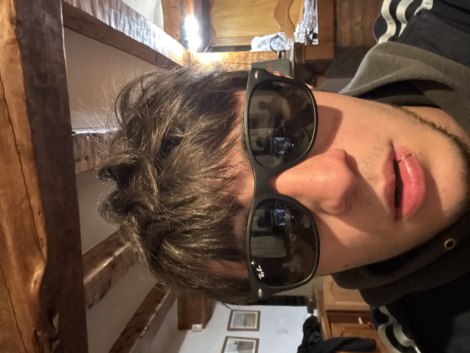 A person wearing sunglasses and a gray hoodie poses for a close-up shot. The sunglasses reflect the interior of a room. The person has short, messy dark hair, fair skin, and a lip piercing. The background features wooden beams and furniture, suggesting an older building interior. Two framed pictures can be seen on the wall.