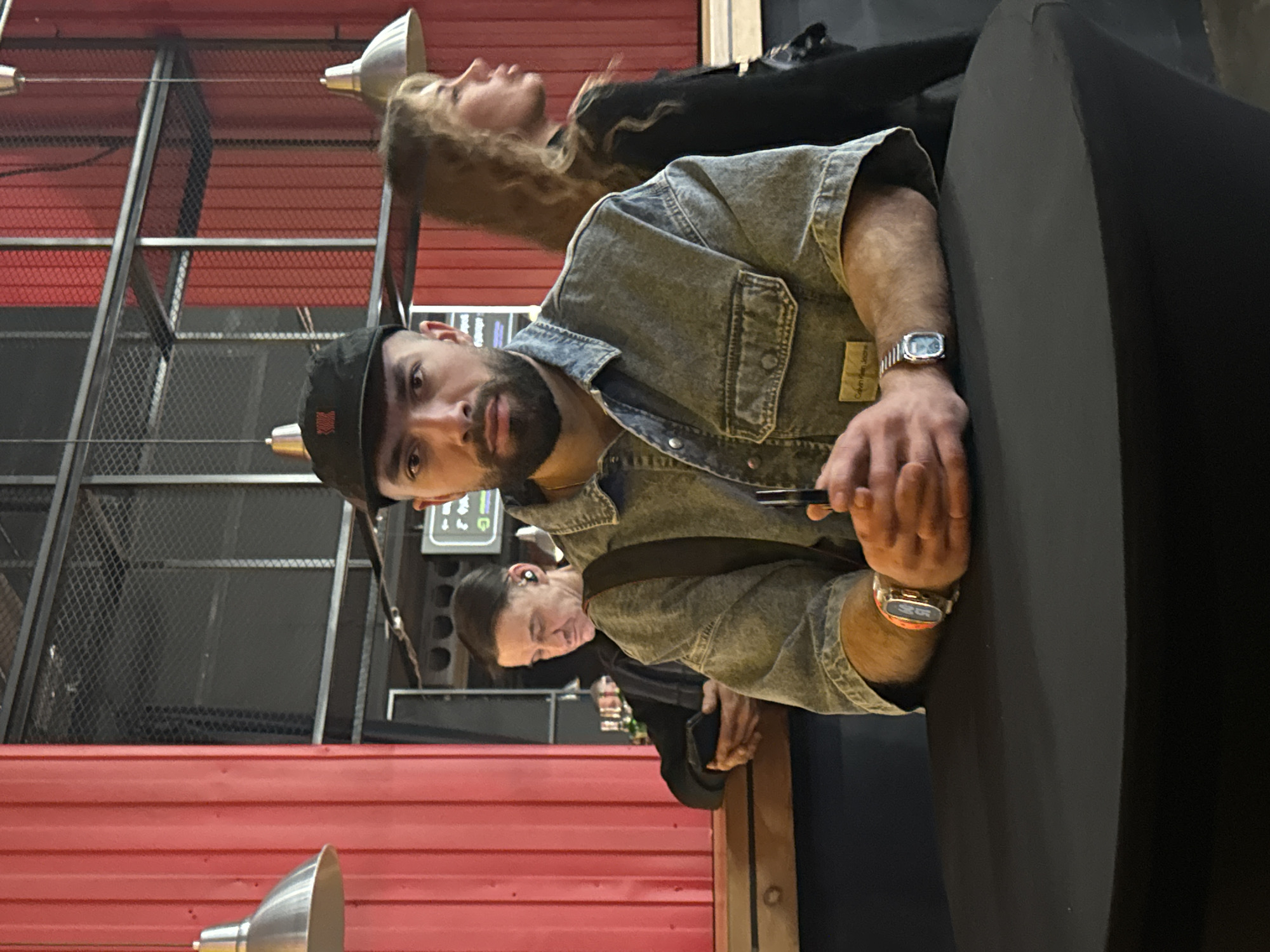 Here is a description of the image:
The image shows three people, primarily focusing on a man in the foreground seated at what appears to be a bar or counter, dressed in a denim jacket and wearing a black baseball cap. He has a beard and is looking towards the camera. There's a wristwatch on his left wrist.
Behind him, to the right, there is a woman with blonde curly hair. Behind him, to the left, there is a man looking to the left, with an earbud in his ear. The background includes red corrugated metal panels and industrial-style lighting fixtures. The location appears to be indoors, possibly a restaurant or bar. The Calvin Klein Jeans logo is visible on the man's sleeve. This scene is not necessarily located in Khimki, Russia; no information present suggests that.