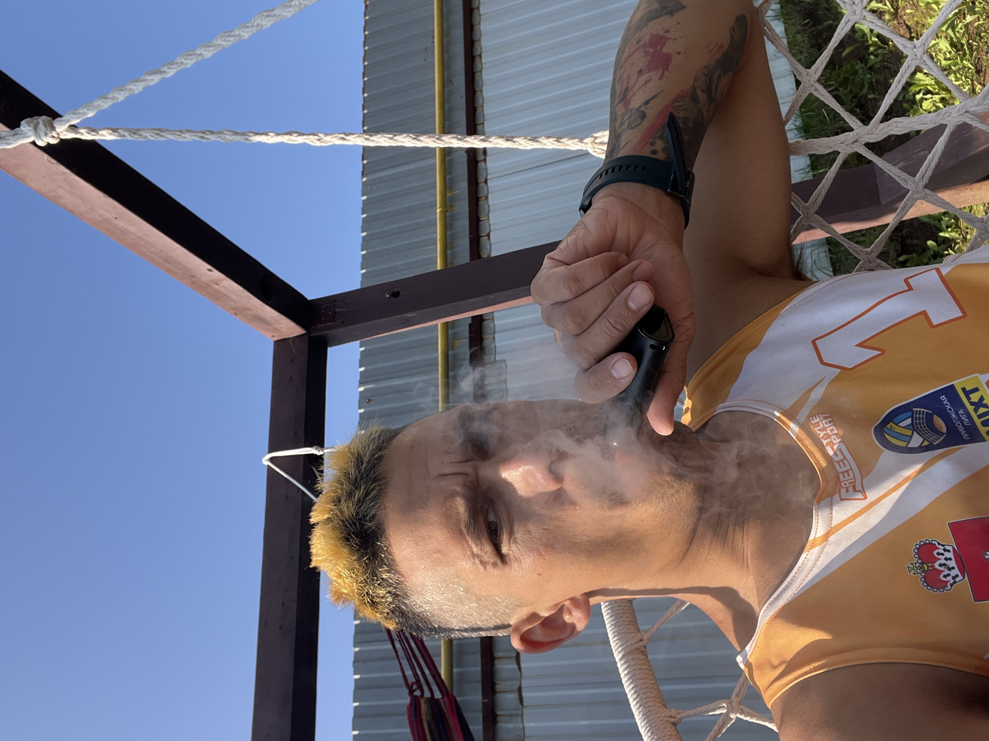 A man with bleached blonde hair, wearing a white and orange sports jersey, is seated in a hammock chair outdoors. He is exhaling smoke from an electronic cigarette. Behind him, a corrugated metal fence and a wooden structure supporting the hammock are visible. The scene appears to be a residential outdoor area, possibly in the city of Chaikovsky, Russia, given the implied location context from the provided metadata. The man has tattoos on his arm and is wearing a dark smartwatch. The sky above is clear and blue.