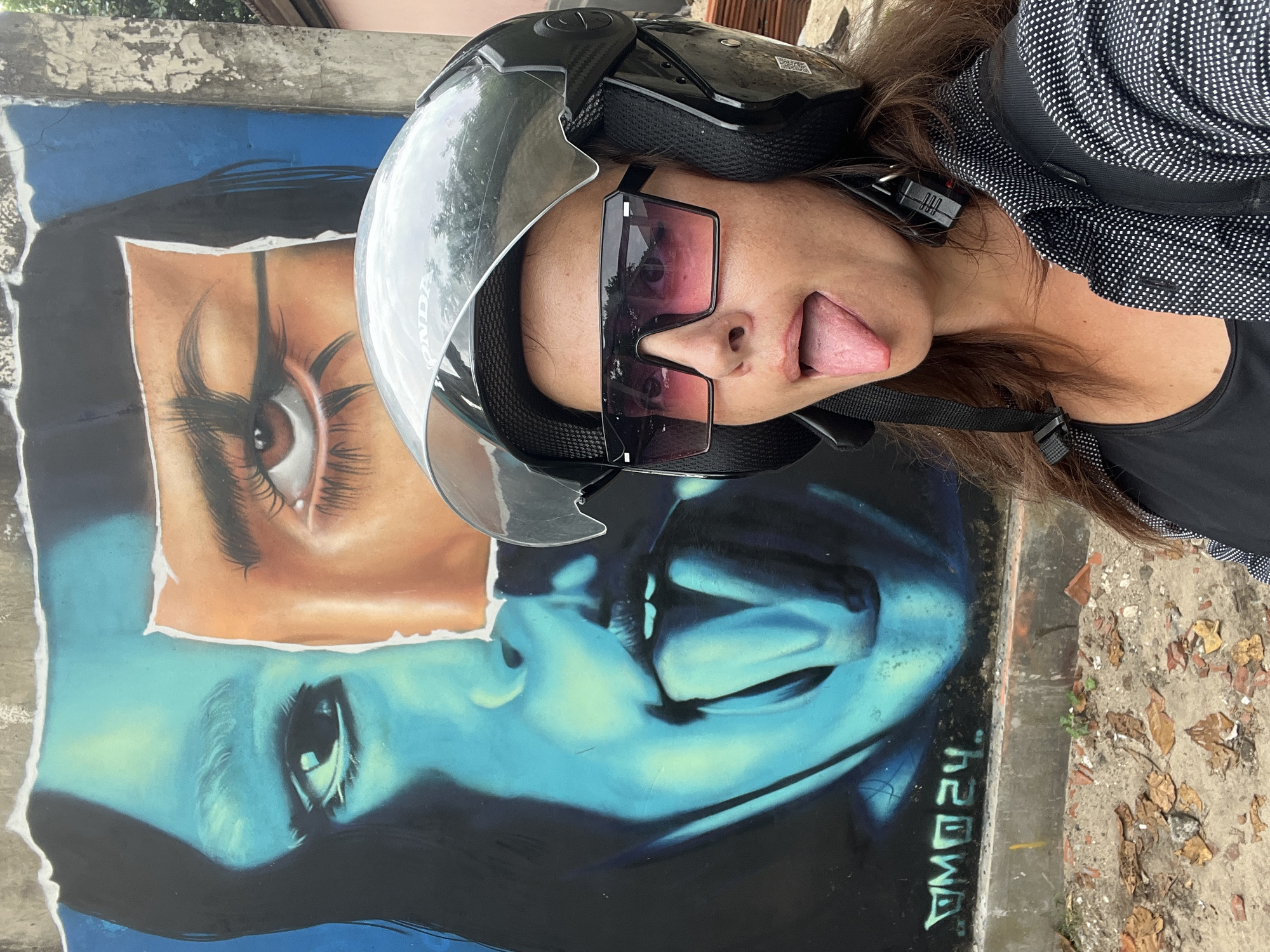 A woman wearing a black helmet and sunglasses with pink-tinted lenses sticks her tongue out at the camera. She is in the foreground, and behind her is a large mural of a woman's face. The mural is split down the middle, with one side depicted in cool blue tones and the other in warmer orange and brown tones. The woman in the mural has one eye open and the other closed, and her tongue is also sticking out. The scene appears to be outdoors, possibly in a city, with bricks and debris visible on the ground. The mural has the signature "DM024" in the lower left corner.