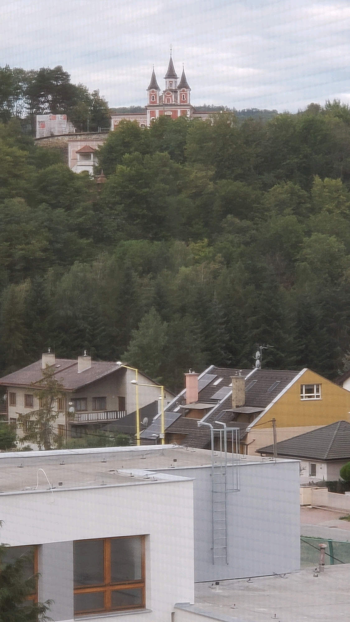 The image shows a cityscape, likely in Prešov, Slovakia, with several buildings and a dense forest on a hill. In the background, a prominent red and white building with multiple spires stands atop the hill, suggesting a religious or historical structure. In the middle ground, there are residential buildings with tiled roofs, chimneys, and solar panels visible. In the foreground, a modern white building with large windows and a metal exterior ladder is depicted. The sky is overcast, indicating a cloudy day. The overall scene is urban with natural elements, showing a blend of architectural styles and the surrounding environment.