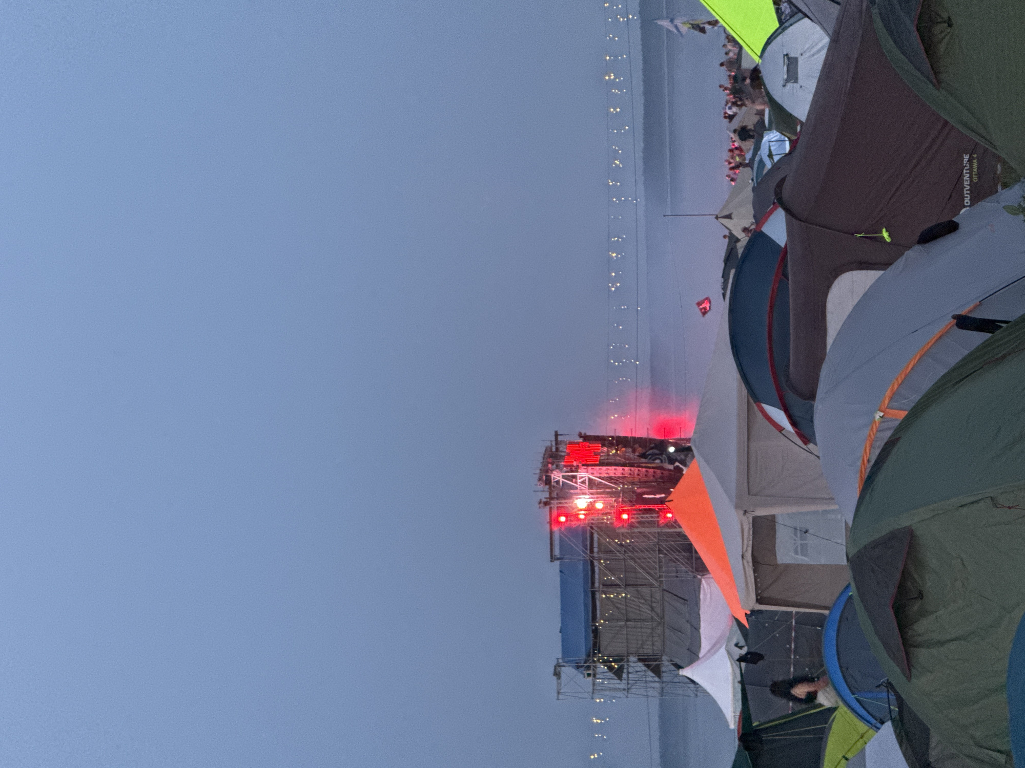 The image depicts a campsite with numerous tents set up in front of a stage with bright red lights. The sky is a pale blue, suggesting either early morning or late evening. In the background, there is a body of water, possibly a lake or the sea, with a faint outline of land on the horizon. String lights are visible in the distance, adding to the festival atmosphere. A few people are seen walking around, but they are not the primary focus of the image. The tents are of various colors and sizes, indicating that this is likely a large event or festival.