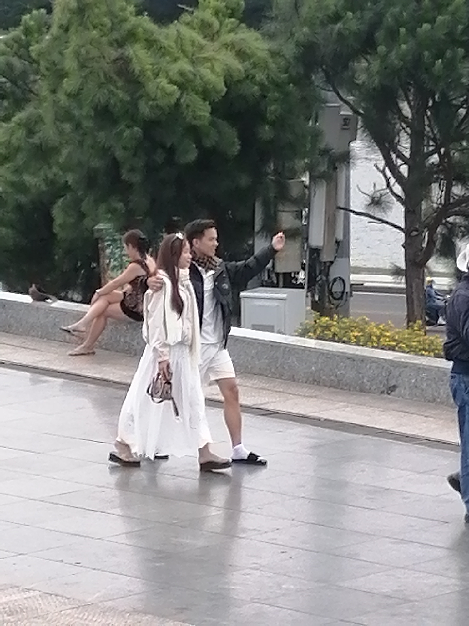 The image depicts a couple strolling along a paved walkway in Da Lat, Vietnam. The woman, in a white, flowing dress and sandals, carries a handbag. The man, in shorts and a black jacket with a patterned scarf, has his arm around her. In the background, another woman is seated on a bench, and trees line the area. A body of water is visible beyond a low wall. The overall scene is one of casual outdoor activity in a city environment.