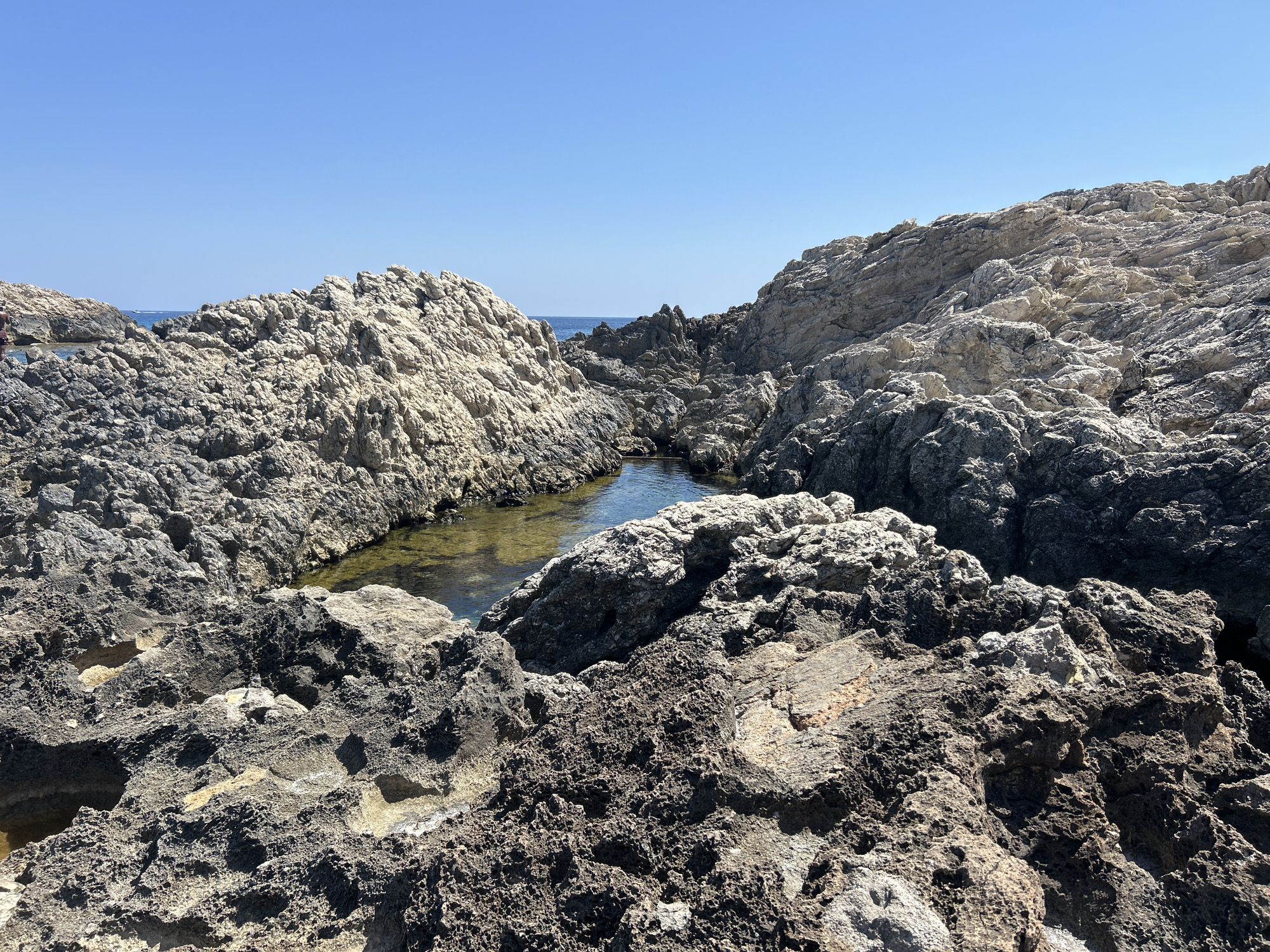 The image shows a natural rock formation along the coast of Cala Lliteres, Spain. The foreground is dominated by rough, textured rocks, predominantly dark gray and brown, with some lighter, sandy-colored sections. These rocks create a rugged and uneven landscape. In the mid-ground, a small pool of clear, shallow water is visible, nestled between the rock formations. The water reflects the bright blue sky. Beyond the pool, more rock formations continue, with a darker, more jagged appearance. The horizon shows the vast expanse of the sea under a clear, bright blue sky. The scene is bathed in direct sunlight, casting shadows on the rocks. There are no visible people in the immediate foreground or mid-ground, though a hint of a person in dark swimwear can be seen on the far left edge, near the water's edge. The overall environment is natural and undeveloped, characteristic of a coastal area with rocky shores.