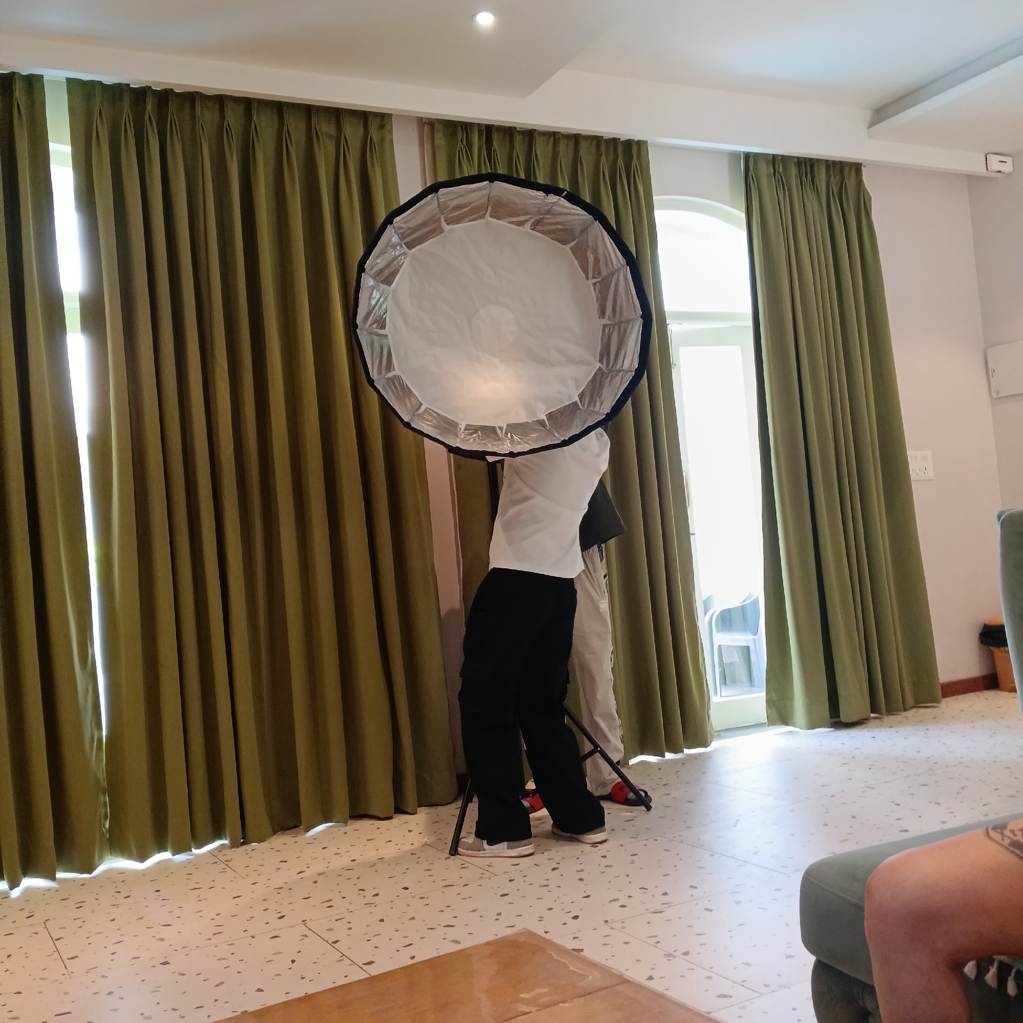 A medium indoor shot in Calangute, India, depicts two individuals standing behind a large, circular reflector on a tripod. The person in the foreground wears a white shirt and black pants, while the second person is dressed in a white shirt and light tan pants. The reflector is held up by the two individuals and is partially obstructing them from view. Behind the people and reflector are olive green curtains with a white window, and to the right of the window is a wall with a light switch. In the foreground, parts of a wooden table and the side of a green sofa are visible. The floor is covered with cream tiles with light brown speckles. A ceiling light fixture is visible in the upper middle area of the picture.