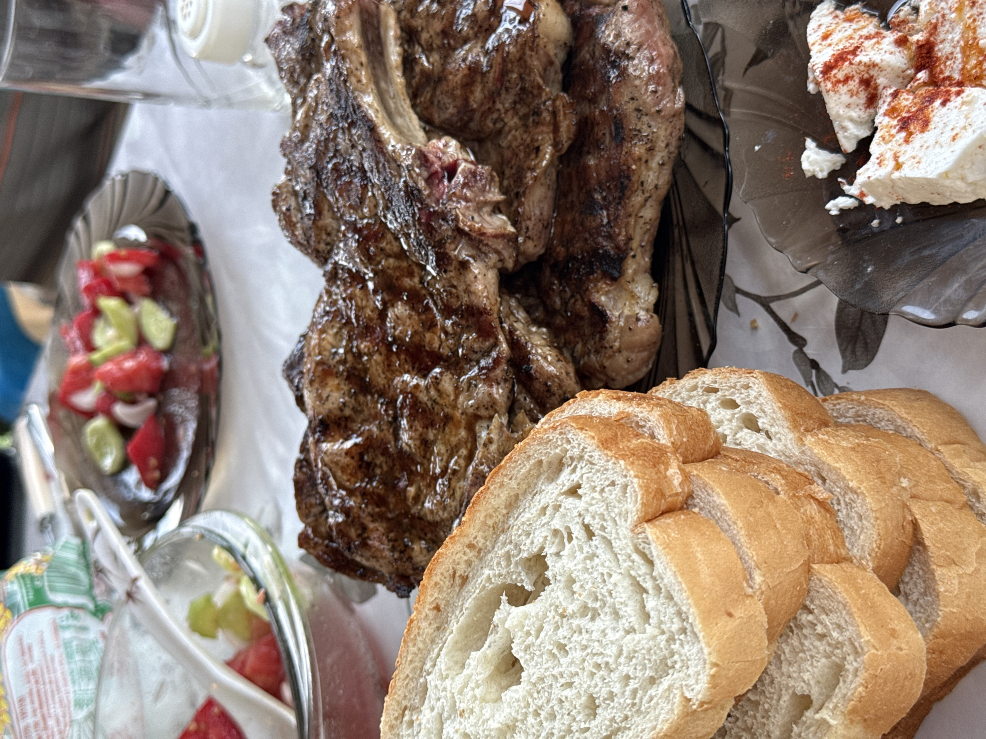 This is an eye-level, close-up, outdoor, still life photograph taken in Ravadinovo, Bulgaria, depicting a meal spread on a white tablecloth. The foreground is dominated by a stack of several thick slices of white bread, with a crusty, golden-brown exterior and a soft, airy interior. To the right of the bread, a pile of grilled steaks, appearing to be ribeye, is presented on a dark, translucent plate. The steaks are well-seared, with visible grill marks and a glistening, juicy appearance. In the background, to the left, a glass bowl contains a chopped salad of tomatoes and cucumbers. Further to the right, another dark, translucent plate holds chunks of white cheese, possibly feta, generously sprinkled with paprika. A clear glass and a white salt shaker are also visible in the background. The overall scene suggests a casual outdoor dining experience.