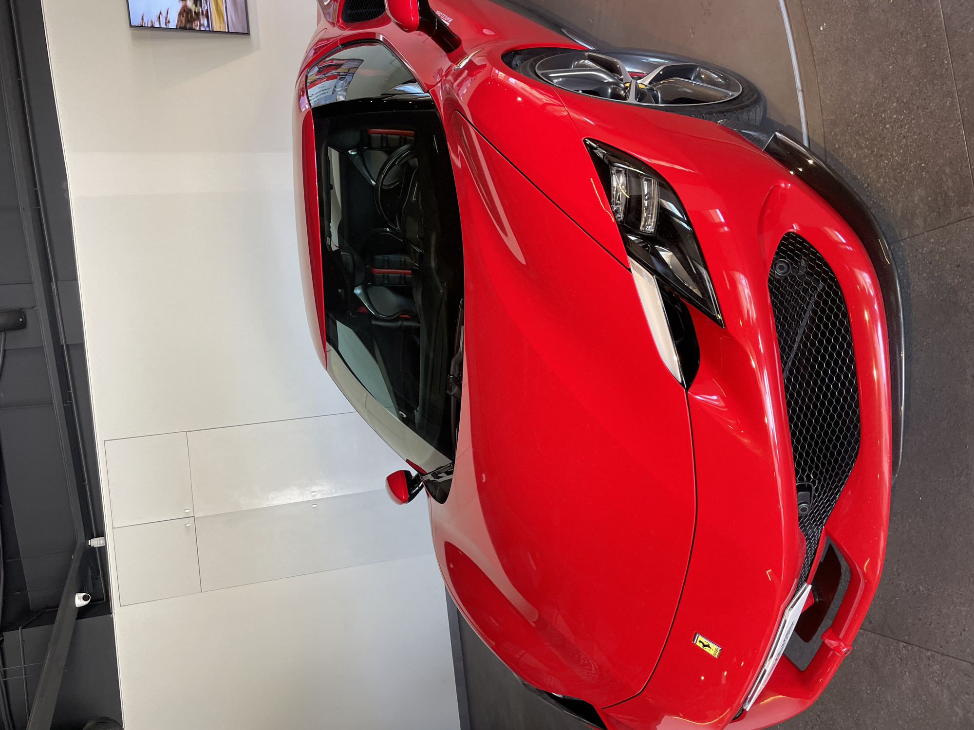 A high-angle shot captures a vibrant red Ferrari sports car on display, likely within a showroom in Barcelona, Spain. The car occupies the majority of the frame, angled diagonally with its front and passenger side prominent. Its sleek, aerodynamic design is highlighted by the bright red paintwork, contrasting with black accents such as the intricate front grille, housing a camera, and the modern headlights featuring distinctive LED elements.
Through the passenger side window, a glimpse of the car's interior reveals a black cabin with sports seats and a steering wheel, accented with subtle red detailing. A small, yellow Ferrari emblem with the iconic prancing horse is visible on the front bumper. On the side bodywork near the windshield, a partial word, possibly "MONOTEQUE" or similar, is visible on a sticker.
The background consists of clean, white walls that reflect some light, enhancing the car's shine. A wall-mounted screen in the upper background displays an image, though its content is not clearly decipherable. The floor beneath the car is dark, possibly concrete or polished asphalt, suggesting a showroom or exhibition space. Overhead, white ceiling panels and a security camera are visible. The overall impression is one of a pristine and controlled environment designed to showcase the luxury vehicle. No people or specific activities are depicted, focusing solely on the stationary car.