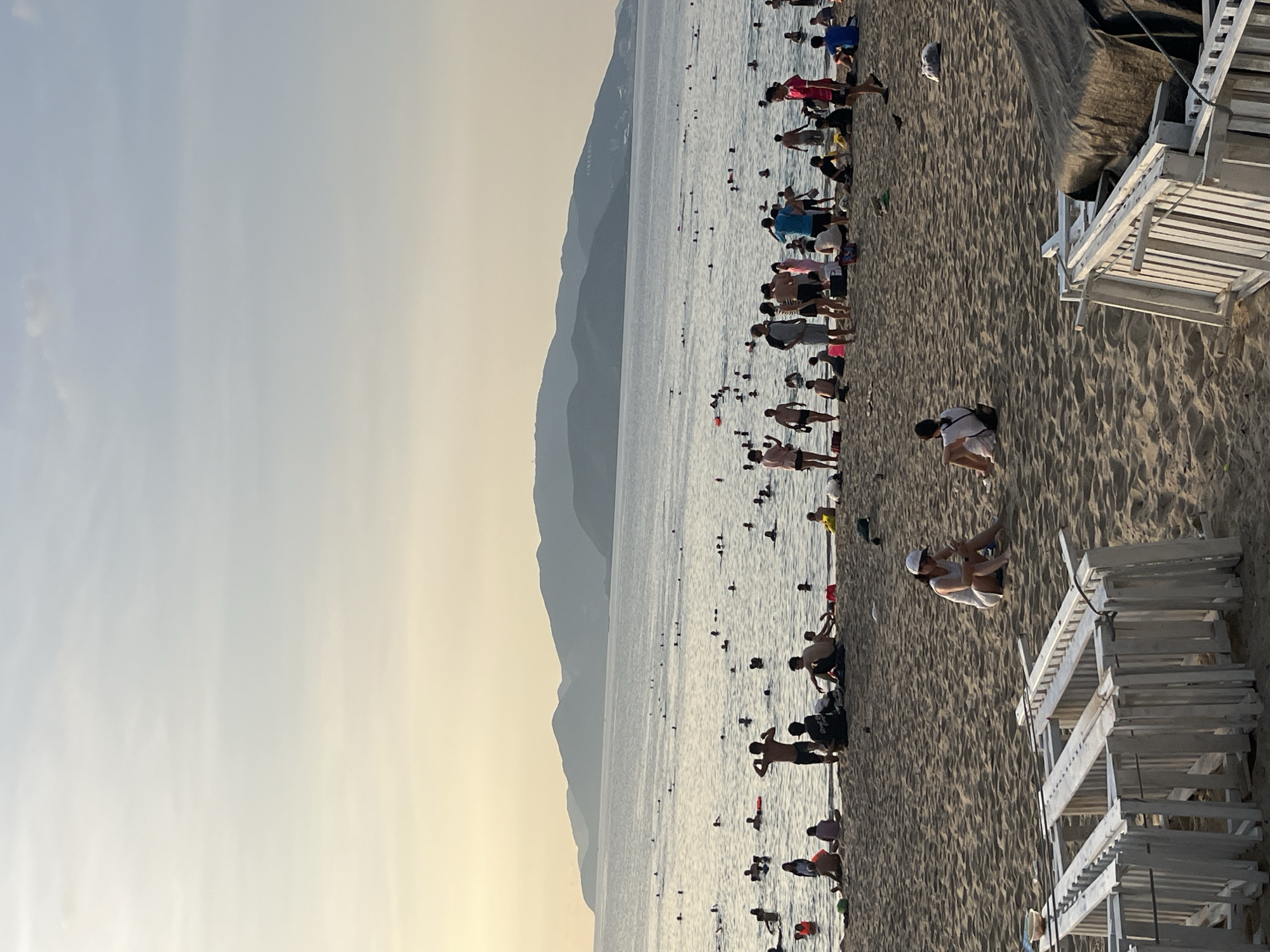 The image depicts a beach in Nha Trang Ward, Vietnam. A large group of people is present, both in the water and on the sand. The water is calm, with numerous individuals swimming or standing in it. On the sandy beach, there are people walking, sitting, and interacting. Mountains are visible in the background, lending a scenic element to the scene. Several white beach chairs are stacked on the right side. The sky is pale and clear, suggesting it may be early morning or late afternoon.
