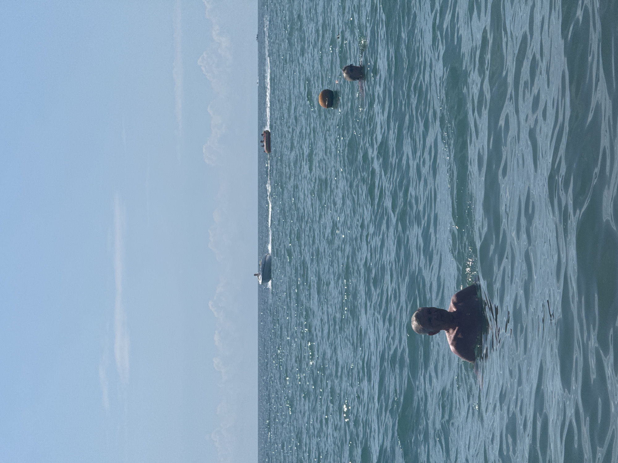 A man with short gray hair is swimming in the ocean in Sochi, Russia, looking towards the camera. The water is choppy and a bright blue. In the distance, a speedboat is visible, and further out, a tube ride with people on it is being pulled by a boat. There are a couple of buoys in the water, one orange and one smaller and darker. The sky is a clear light blue with a few scattered white clouds. The overall scene depicts a sunny day at the beach with various water activities.
