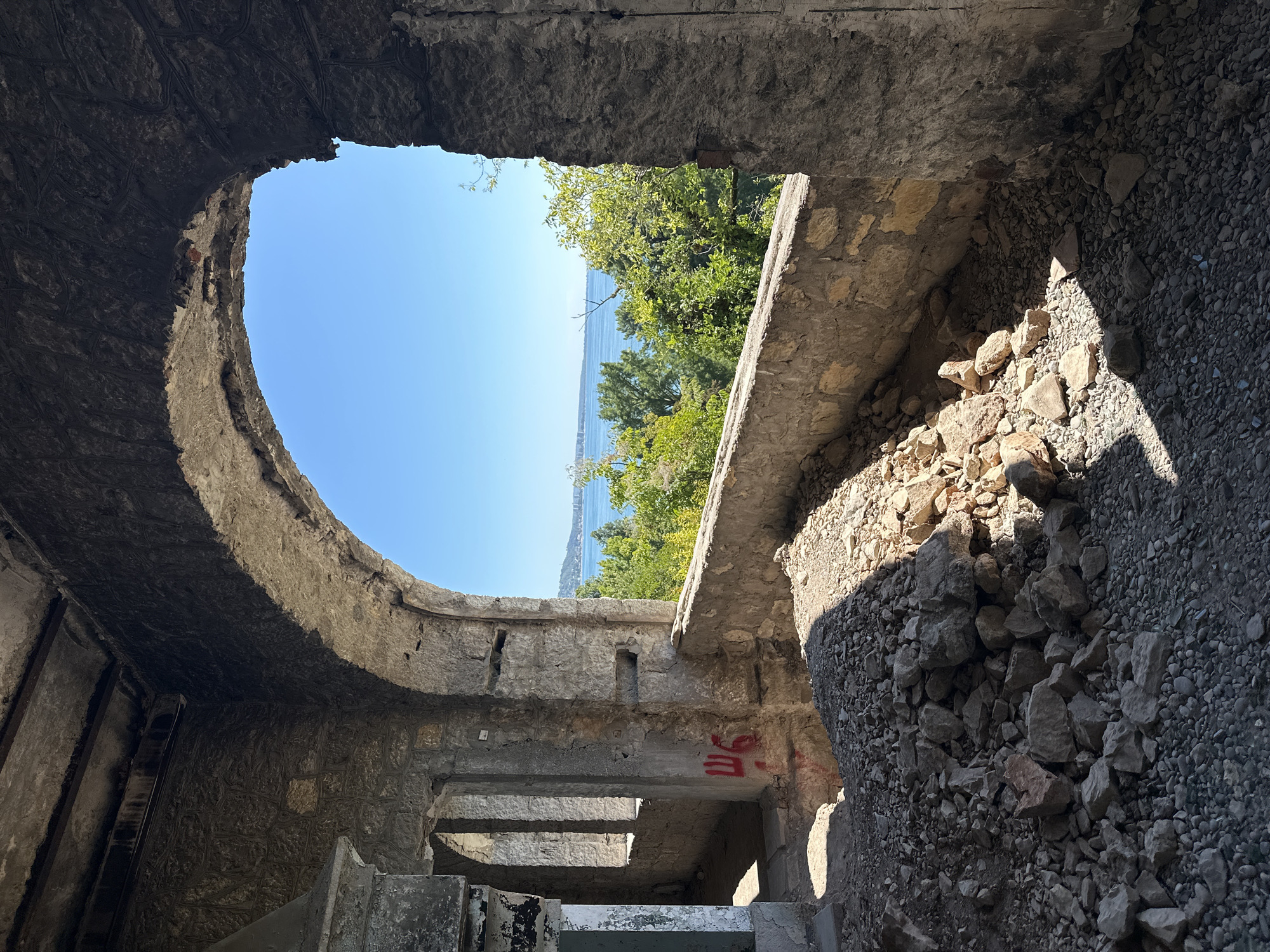 The image depicts the interior of a dilapidated stone structure, likely an abandoned building, overlooking a body of water. The foreground is filled with rubble and loose stones. An arched opening frames a view of a calm, blue body of water, likely a sea or large lake, under a clear blue sky. Lush green trees and vegetation are visible between the structure and the water, with a distant coastline and some buildings suggesting an inhabited area. The architecture of the interior suggests a historical or possibly ancient construction, with rough-hewn stone walls and evidence of decay. Red graffiti, appearing to be Cyrillic characters, is visible on one of the interior walls. The lighting suggests a bright, sunny day, with strong shadows cast by the interior architecture onto the rubble. The scene evokes a sense of abandonment and the passage of time, contrasted with the vibrant natural landscape beyond. The location is described as Abaata, Abkhazia.