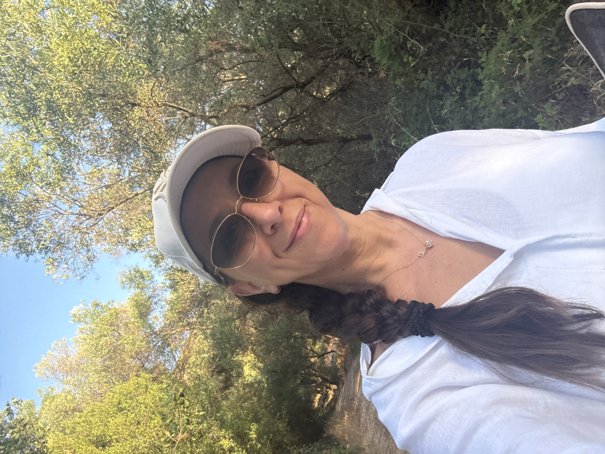 A woman is taking a selfie while walking outdoors in Makrades, Greece. She is wearing a white button-down shirt, a white baseball cap, and sunglasses. Her hair is in a braid and she is wearing a delicate necklace. She is smiling at the camera. The background shows trees and a path, suggesting a natural, outdoor setting. The sun is shining, casting a warm light on the scene.
