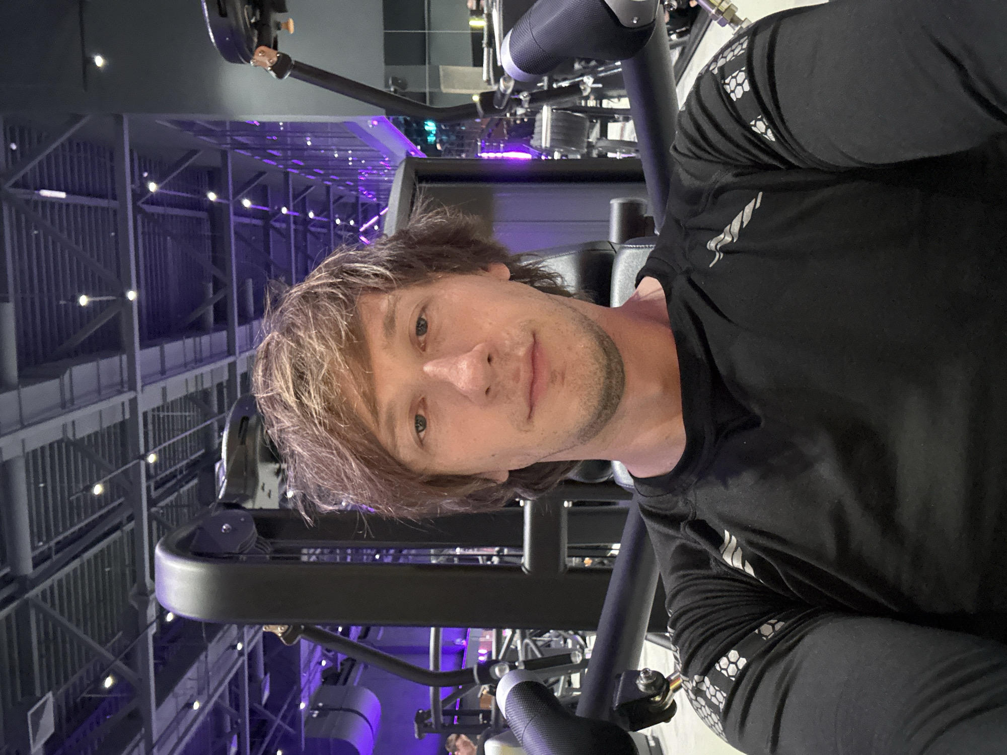 A man is taking a selfie in a gym. He is wearing a black long-sleeved shirt with silver geometric patterns on the sleeves. The gym has purple ambient lighting and industrial-style architecture with exposed beams and metal structures. Various gym equipment, including a large black machine, is visible in the background. The man has medium-length, light brown hair and appears to be looking directly at the camera. The image is taken from a slightly low angle, and the overall mood seems casual and focused on fitness. The time of day is not clearly indicated, but the indoor lighting suggests it could be anytime. There is no visible text in the image.