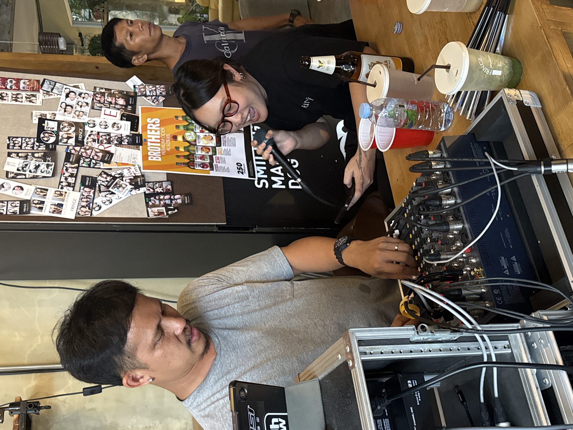 In Chiang Mai City Municipality, Thailand, a man wearing a grey t-shirt and a silver earring is operating audio equipment. He has dark hair and a mustache and is looking down at the console. Beside him, a woman with glasses and a black t-shirt is holding a microphone and appears to be singing. She has dark hair and is looking to her left. In the background, a man in a dark t-shirt stands and looks to the right. The scene is set in what appears to be a bar or cafe, with a wooden counter in the foreground displaying drinks and audio equipment. A collage of photos is visible on the wall behind them, along with a poster advertising "BROTHERS PREMIUM CIDER".