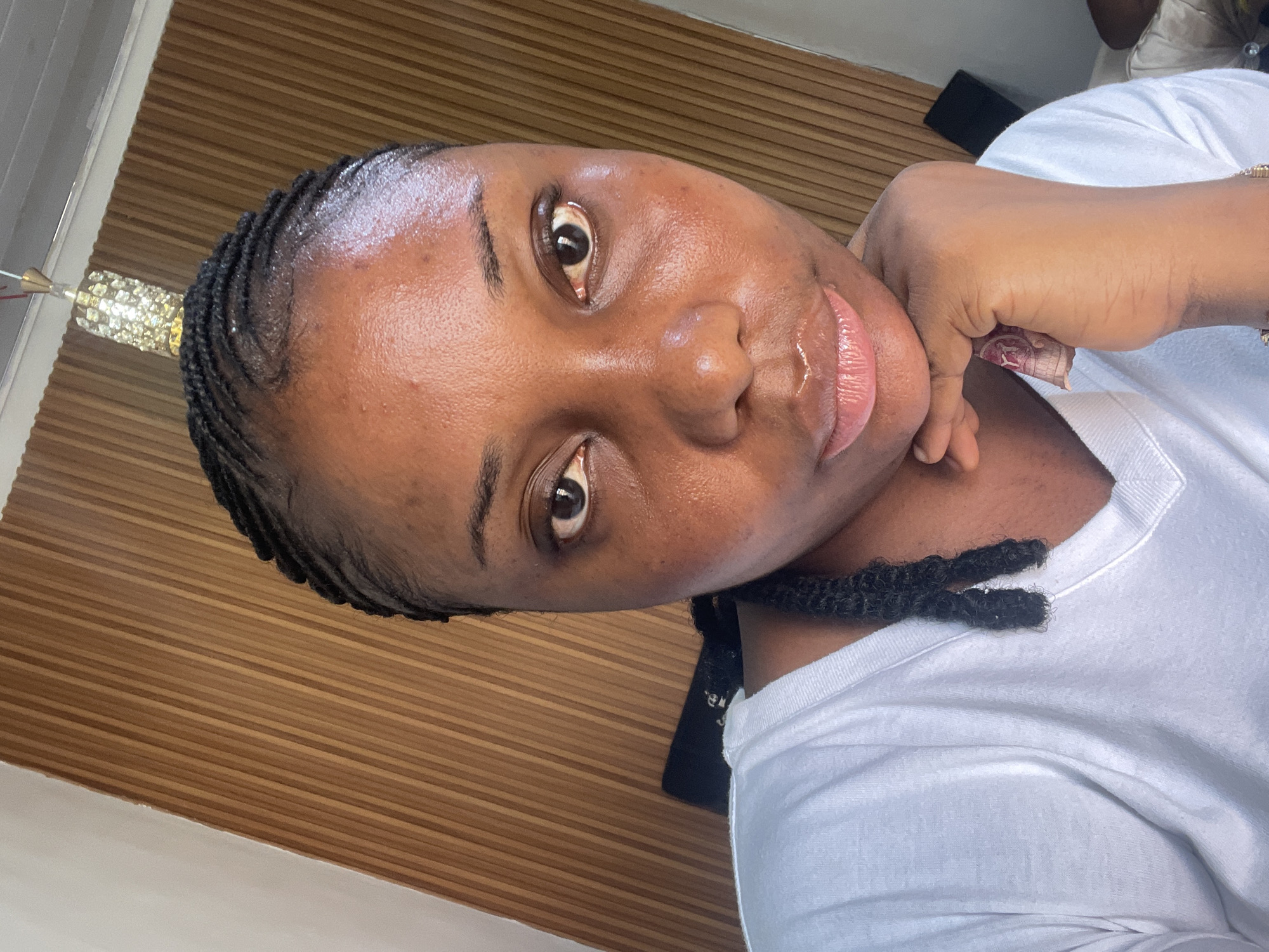 A medium close-up shows a Black woman looking directly at the camera. Her face is shiny. She has neatly braided hair, and she wears a white v-neck t-shirt. Her right hand, with a gold-toned bracelet, is raised to her face, supporting her chin. A partial object emitting light is located at the top left. The background features a wall with a wood paneling design. The scene is likely indoor, with good lighting conditions. The image was taken in Abuja, Nigeria.