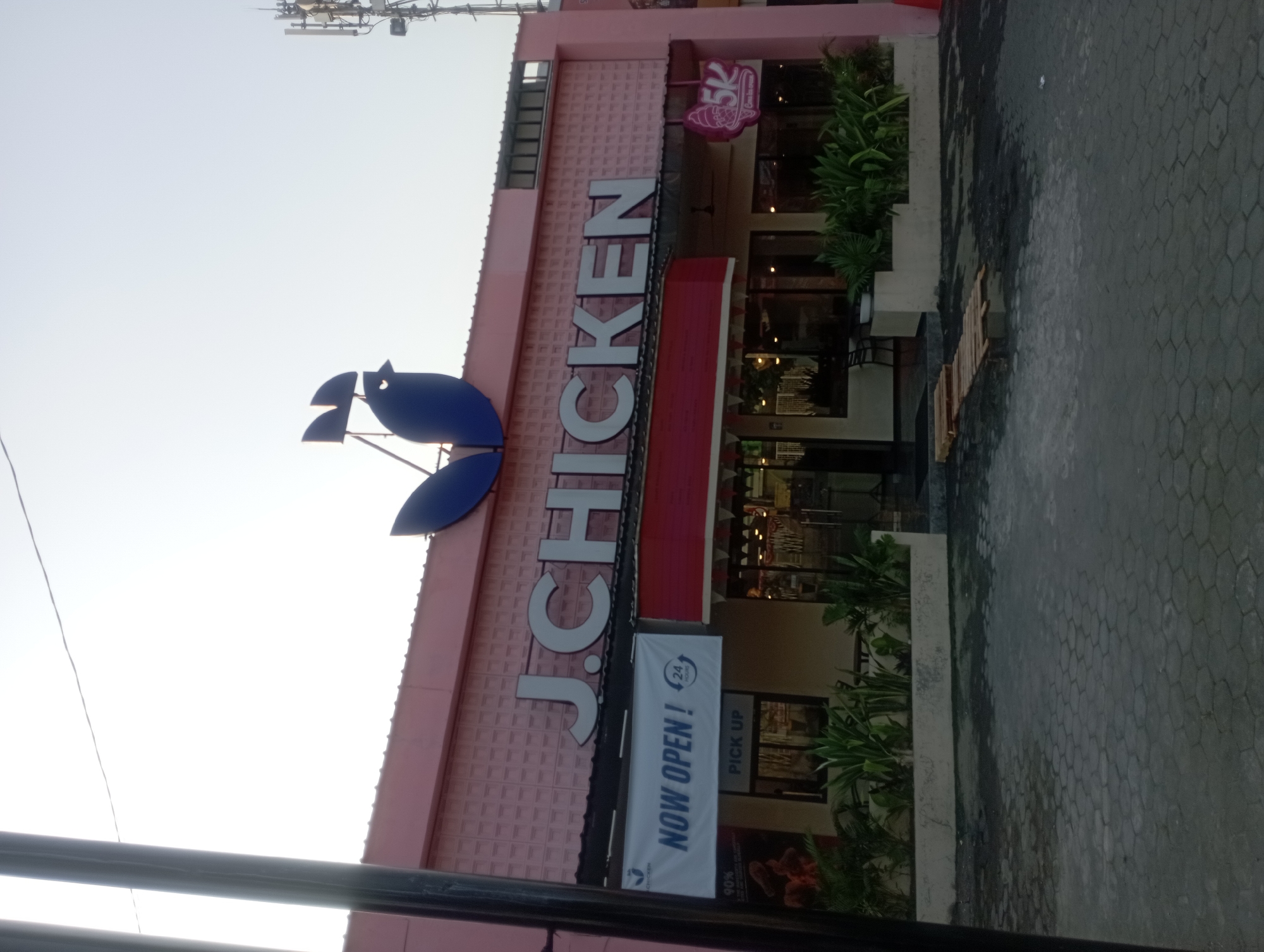 The image shows the exterior of a "J. CHICKEN" restaurant in Yogyakarta, Indonesia. The building is painted pink and has a large sign with the restaurant's name and a blue bird logo. A banner across the front reads "NOW OPEN! 24 HOURS". There is a "PICK UP" sign visible. The foreground shows a paved area with hexagonal tiles. Lush green plants are arranged in planters in front of the building. A cellular tower is visible in the background to the right. The lighting suggests it is daytime, likely in the late afternoon or early morning due to the soft, diffused light. There are no people visible in the scene.