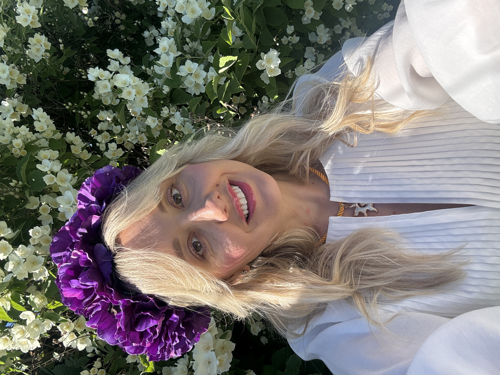 A young woman with blonde hair is lying down outdoors, surrounded by white flowers and greenery. She is wearing a white, pleated shirt and a large, vibrant purple floral crown around her head. The woman is smiling widely, showing her teeth and pink lipstick. A delicate gold necklace with a silver pendant is visible around her neck. The scene appears to be set during the daytime, likely in the summer due to the abundance of blooming flowers. The lighting suggests a sunny day, casting soft shadows and highlighting the woman's features and the textures of her clothing and the flowers. The overall impression is one of joy and natural beauty. The location is Carnikava, Latvia.