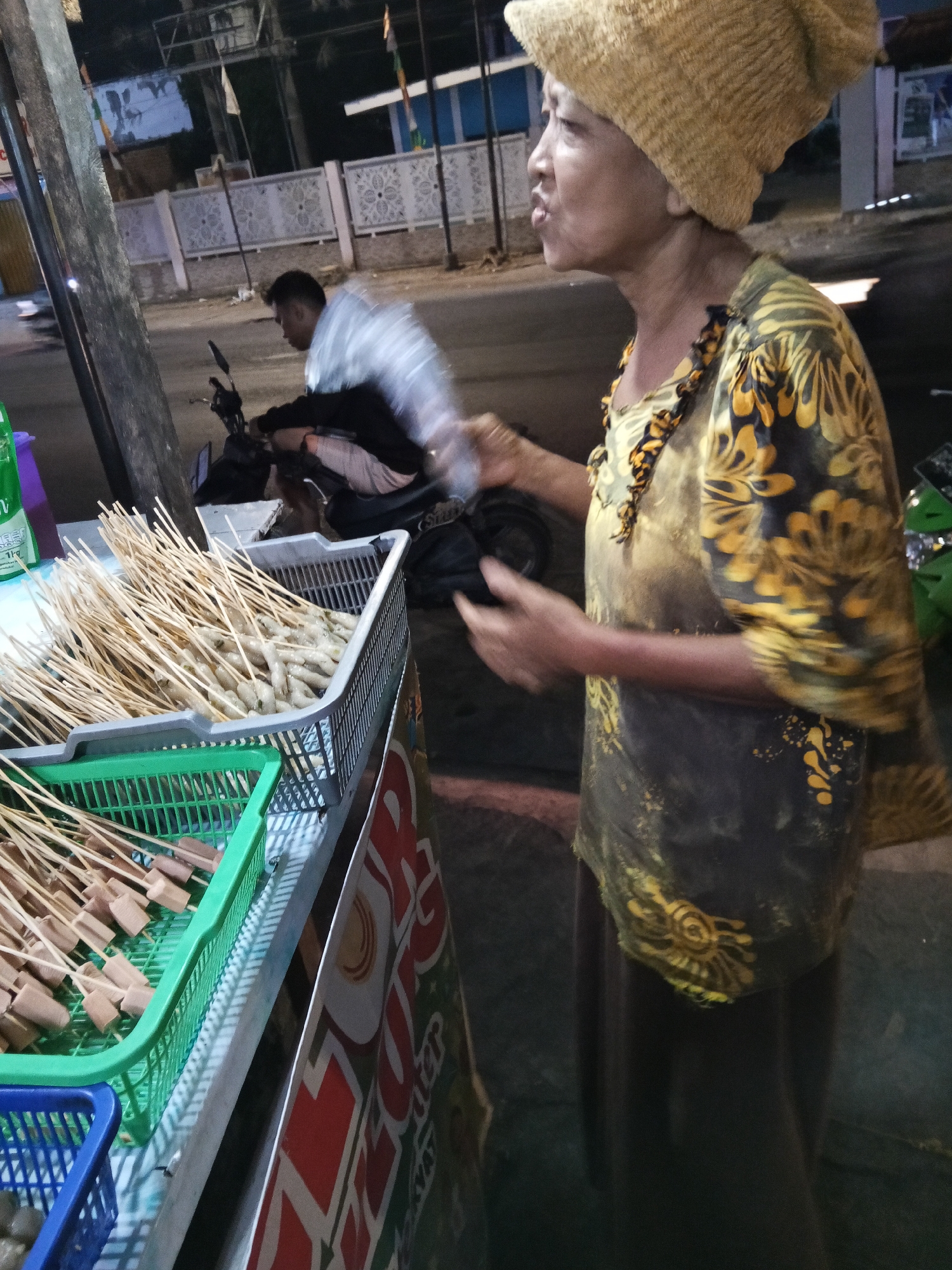 In a nighttime urban scene, likely a street food vendor in Lubuklinggau, Indonesia, a woman wearing a yellow head covering and a dark shirt with yellow floral patterns is captured in profile, speaking. She stands beside a food cart laden with skewers of raw ingredients. To her left, a basket holds what appear to be sausages on skewers, and another basket further back contains skewers of what might be fish balls. A motorcycle is visible in the background, with a person on it whose clothing is blurred, suggesting movement. The environment includes a road, sidewalk, and a building with decorative fencing. The lighting suggests the presence of streetlights or artificial illumination from the vendor's stall.