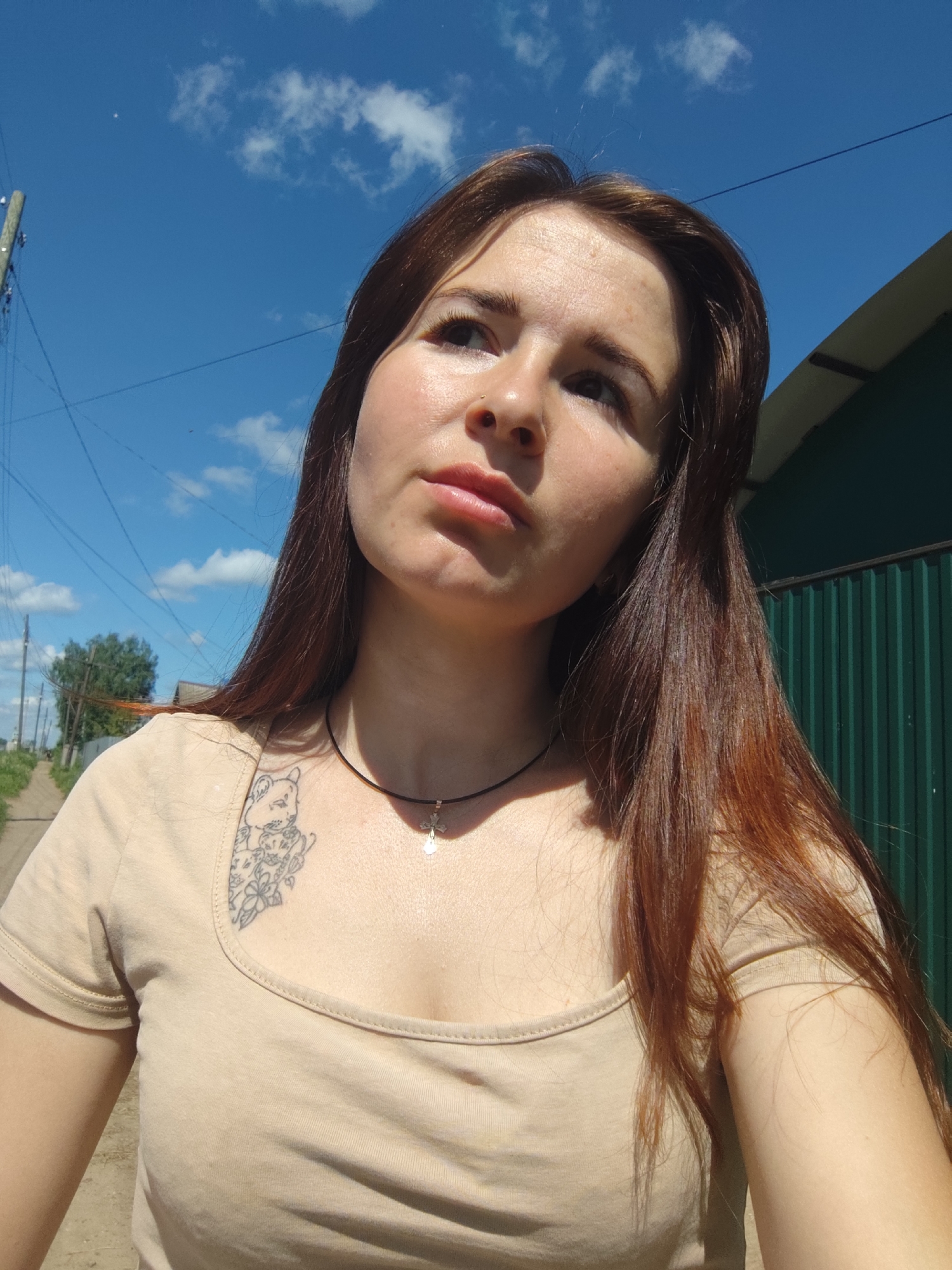 A young woman with long, brown hair stands outdoors, looking upwards. She is wearing a beige t-shirt with a tattoo on her upper left chest and a thin necklace with a small pendant. The background features a bright blue sky with scattered clouds, utility poles and wires, a green corrugated metal fence, and a dirt road leading into the distance. The environment suggests a rural or suburban setting, possibly in the city of Fokino, Russia, given the context.