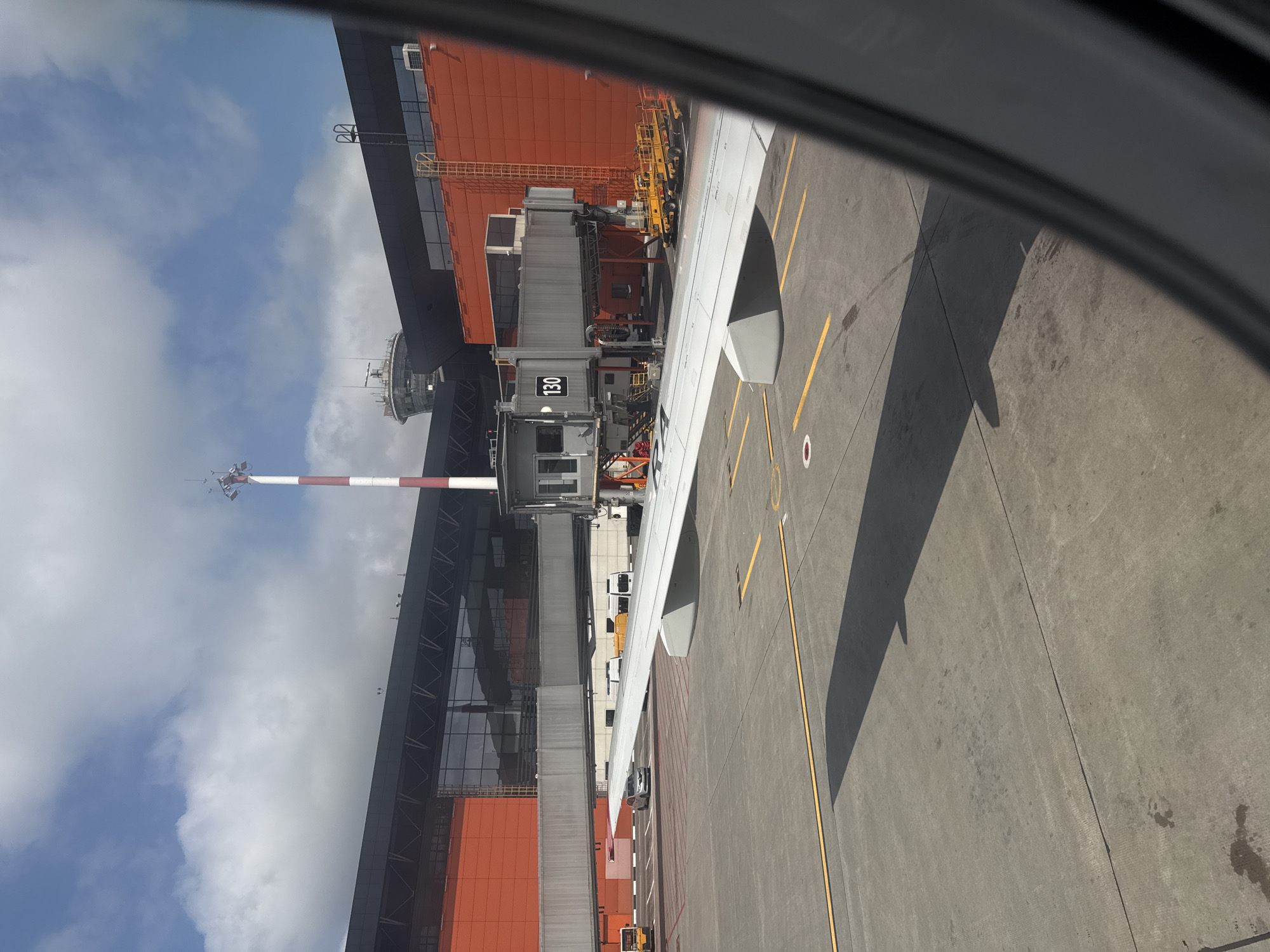 This image shows an airplane wing extending from the bottom left of the frame, casting a shadow on the tarmac. In the background, a modern airport terminal building is visible with a prominent orange facade and large glass windows. A jet bridge, labeled with the number "130," connects to the terminal, and a few airport vehicles are parked on the tarmac. Further in the distance, a control tower with a distinctive circular structure rises against a cloudy blue sky. The scene is characteristic of an airport environment, likely Sheremetyevo International Airport, located in Khimki, Russia, given the architectural style and the presence of a control tower. No people are clearly visible or engaged in any activity.