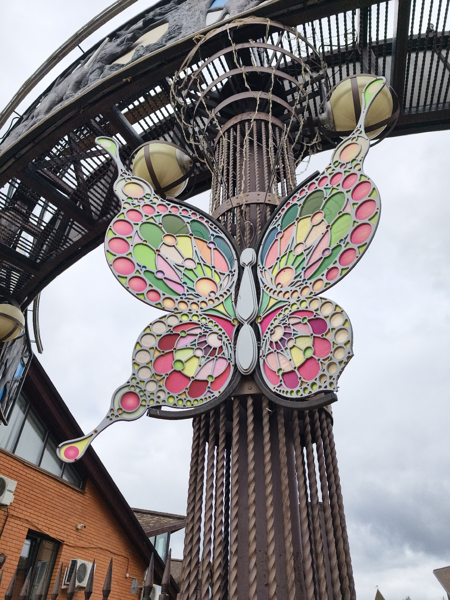 A large, ornate butterfly sculpture, crafted from a metallic frame with vibrant, translucent inserts, is the central subject of the image. The butterfly's wings feature a mosaic of pink, green, yellow, red, and beige hues, arranged in geometric and circular patterns. It is prominently displayed on a tall, dark brown metallic structure composed of twisted, rope-like columns and cylindrical, cage-like sections. Several spherical, light-colored globes, likely decorative lights, are integrated into the upper portion of this support structure.
An overarching metallic arch, also with intricate details including sculptural figures resembling animals or mythical creatures, extends across the upper part of the frame, suggesting the butterfly is part of a larger gate or installation. The sky is overcast and uniformly light grey, indicating a cloudy daytime.
In the lower-left background, a reddish-brown brick building with multiple windows and white air conditioning units is visible. The brand "CHIGO" is discernible on one of the AC units. Dark, pointed elements, possibly from a fence or the base of the sculpture, are present in the foreground. The scene depicts a static architectural or artistic display in what appears to be a public or commercial area, likely in Izhevsk, Russia. No specific activities or people are present.