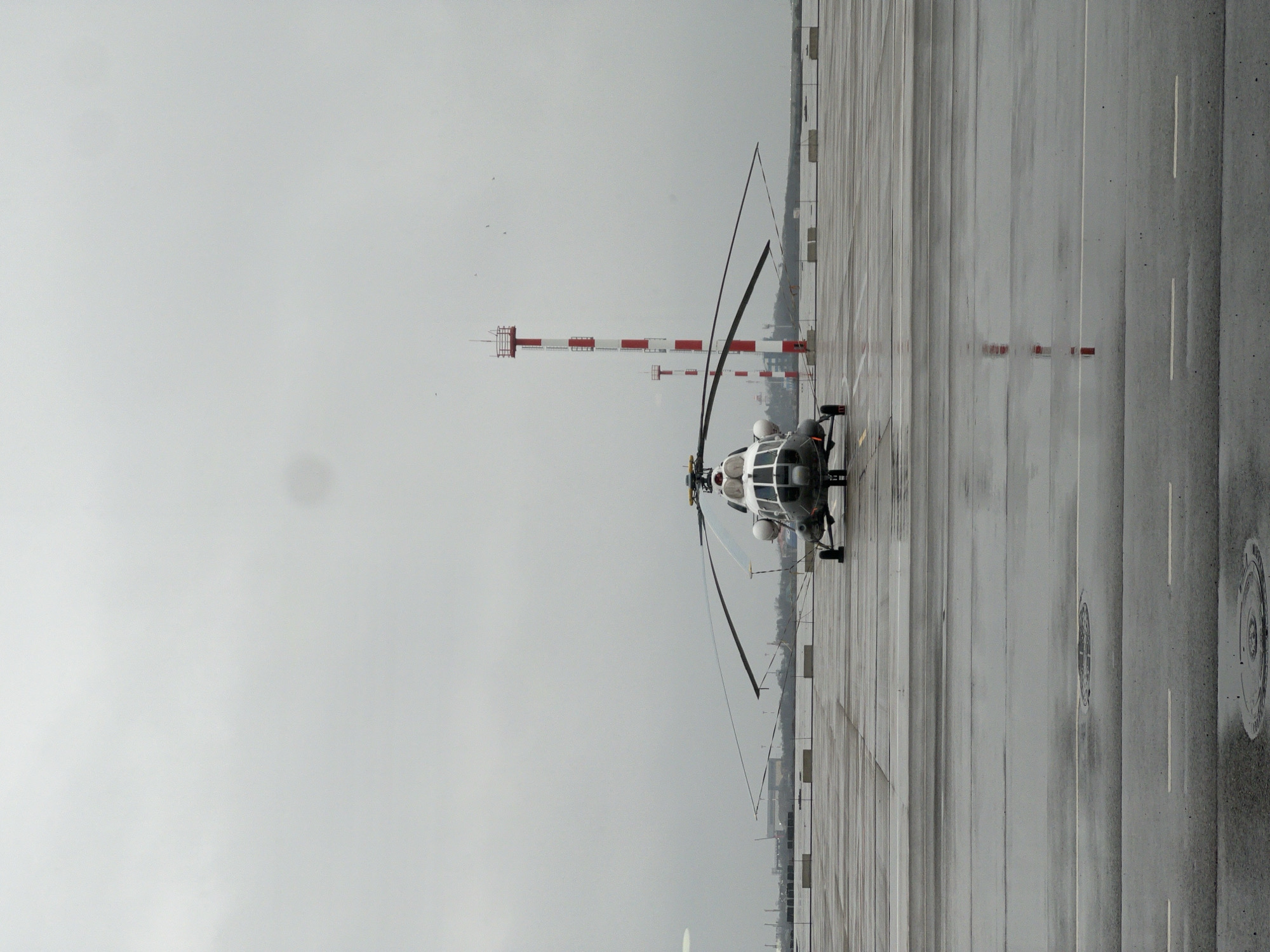 The image depicts a white helicopter with black rotors sitting on a wet concrete tarmac at an airport or airfield. In the background, there are several red and white striped towers, likely part of the airport's navigational equipment. The sky is overcast, suggesting rainy or cloudy weather. The ground is marked with white lines and circular access points, indicative of a functional airport setting. The photograph is taken at ground level, presenting an eye-level view of the helicopter and the surrounding airport infrastructure. While Gorodok is a common place name, given the limited information it is not possible to know if this image is related to the one located in Russia.