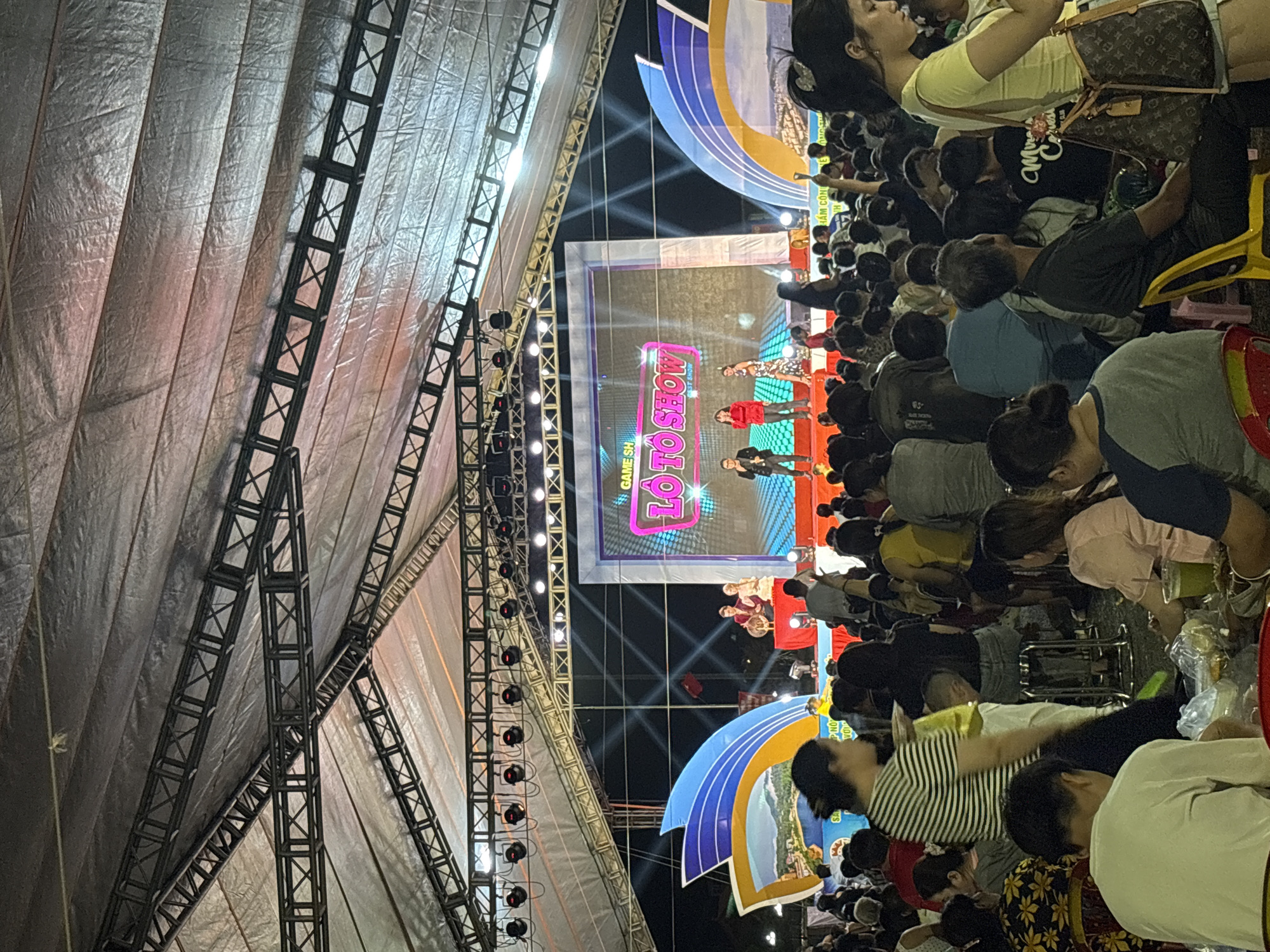 An outdoor stage event is underway under a large, translucent canopy supported by metal trusses. The canopy is lit by numerous stage lights and some ambient lighting, creating a mix of white and colored illumination. A large LED screen is prominently displayed on the stage, showing the words "GAME SHOW LOTO TO SHOW". On the stage, three performers are visible: two men and a woman, dressed in casual attire. One man in a red top and dark pants stands to the left, the other in a dark suit is on the far right, and a woman in a patterned dress is in the center. In front of the stage, a large crowd of people is gathered, seated and standing, facing the performance. They appear to be enjoying the show, with many looking towards the stage. The ground in front of the stage is covered with a red carpet. The overall atmosphere suggests a lively community event or festival.