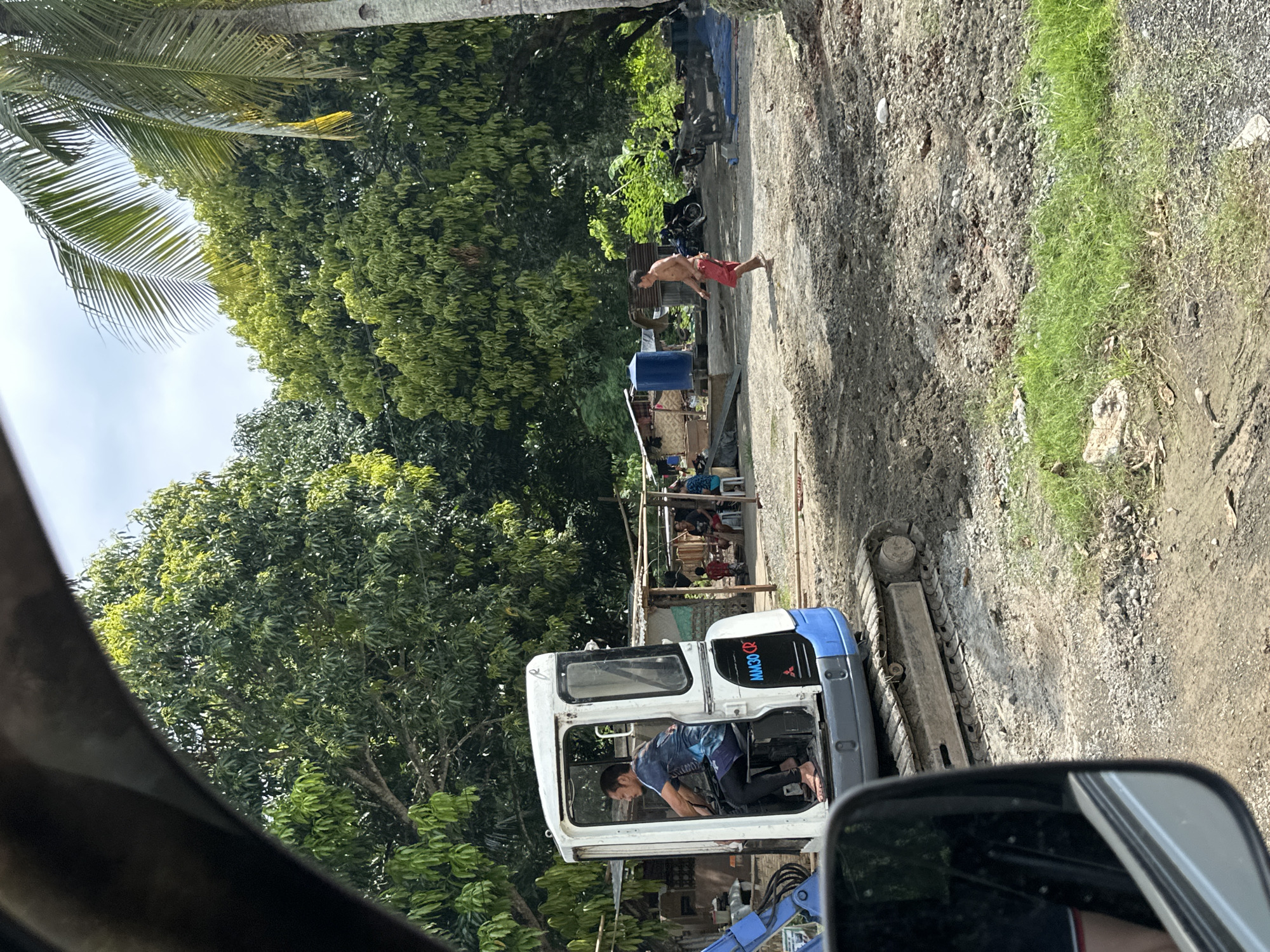 This image captures an outdoor scene in a rural or semi-rural area of Samal, Philippines, viewed from inside a vehicle, indicated by the dark curve of the car's interior at the top and a rearview mirror at the bottom left. The environment is characterized by abundant lush green foliage, including numerous leafy trees and a prominent palm tree on the right. The ground is predominantly dirt and gravel, with patches of green grass.
In the mid-left, a white and blue Mitsubishi MM30 CR mini-excavator with tracks is positioned on the disturbed earth. A man with dark hair and a beard is seated inside the excavator's cab, operating the controls. He is barefoot and wears a blue patterned short-sleeved shirt and dark leggings.
In the mid-ground, a shirtless man in red shorts walks barefoot from left to right across the dirt ground. Further in the background, several rudimentary structures, appearing to be houses or shelters made of wood and corrugated metal, are visible. A blue cylindrical water tank stands near one of these structures. A group of individuals, including a child in a red shirt, are gathered near these structures; some are seated, and one person appears to be sweeping the ground. Two motorcycles are parked in the background to the right of the structures. The ground throughout the scene shows signs of recent disturbance, consistent with excavation work. The lighting indicates daylight, under a partially cloudy sky.