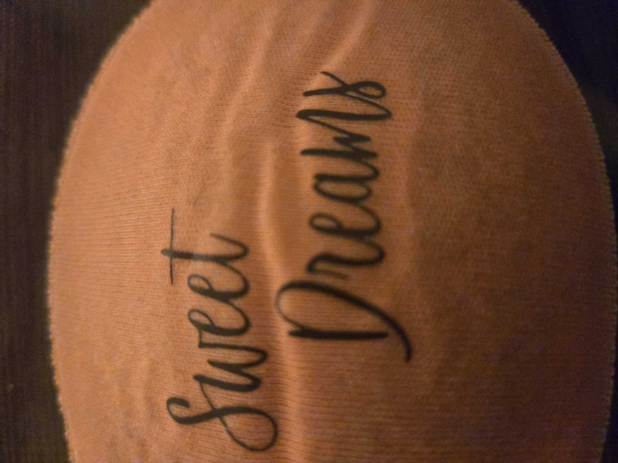 The image presents a close-up, static perspective of a peachy-orange woven textile, likely a pillow, blanket, or soft garment. The fabric is adorned with the dark, cursive phrase "Sweet Dreams," applied to its surface. The textile exhibits a fine, consistent weave pattern and slight surface irregularities, including gentle creases and undulations. The background around the fabric is uniformly dark and indistinct, providing no specific contextual details, architectural features, or unique identifiers. No individuals are present in the scene, and no actions or interactions are depicted. The visual content of the image offers no discernible information to determine its specific geographic location, therefore, its setting cannot be identified as being within the city of Yerevan, Armenia.