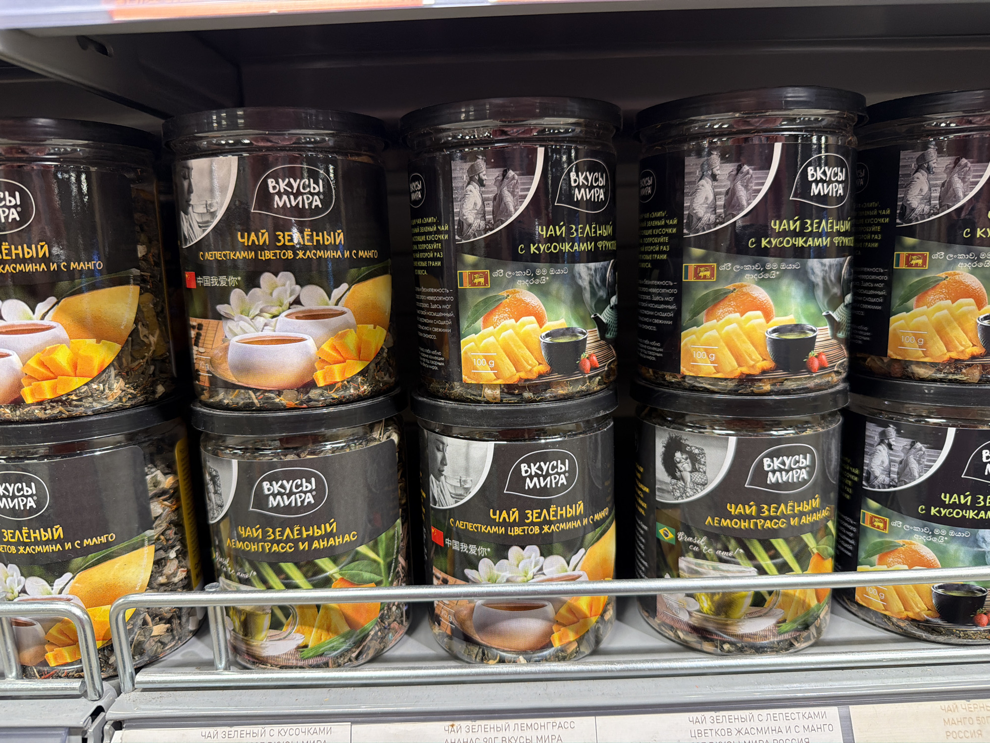 The image shows a close-up of a shelf in a store, likely in Khimki, Russia, based on the text on the shelf tags. The shelf is stocked with multiple identical jars of "Вкусы Мира" (Flavors of the World) green tea in various flavors.
The top row consists of:
- Green tea with jasmine petals and mango. The label features an image of a teacup, white flowers, and mango slices.
- Green tea with fruit pieces. The label depicts what appears to be a couple in Sri Lankan clothing, an orange, and tea.
- Green tea with fruit pieces. The label depicts what appears to be a couple in Sri Lankan clothing, an orange, and tea.
- Green tea with fruit pieces. The label depicts what appears to be a couple in Sri Lankan clothing, an orange, and tea.
The bottom row consists of:
- Green tea with jasmine petals and mango. The label features an image of a teacup, white flowers, and mango slices.
- Green tea with lemongrass and pineapple. The label depicts a woman with a large Afro and a Brazilian flag.
- Green tea with jasmine petals and mango. The label features an image of a teacup, white flowers, and mango slices.
- Green tea with lemongrass and pineapple. The label depicts a woman with a large Afro and a Brazilian flag.
- Green tea with fruit pieces. The label depicts what appears to be a couple in Sri Lankan clothing, an orange, and tea.
The shelf itself is made of metal and has a standard grocery store appearance. Tags below the products indicate the specific names and weights of the tea.