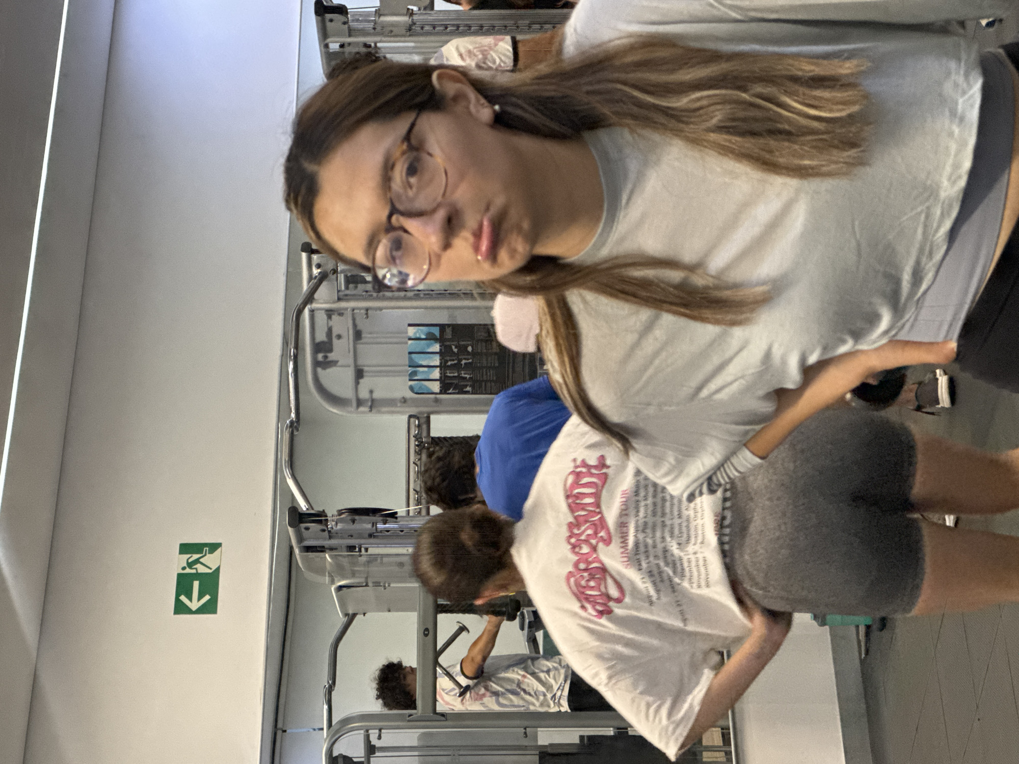 The image captures an indoor scene within a gym or fitness center in San Andrés Cholula, Mexico, featuring several individuals and exercise equipment. The perspective is from a slightly low angle, looking across the room.
In the foreground, a young woman with long, straight brown hair and clear, round-framed eyeglasses is prominent. She wears a plain light gray t-shirt and looks directly at the camera with a somewhat pouting or serious expression.
Behind her, another person is seen from the back, wearing a white t-shirt with a large pink graphic. The text "AEROSMITH SUMMER TOUR" is clearly visible on the shirt, along with a partial list of tour dates and cities such as "August 21," "Phoenix," "San Diego," "Seattle," "Denver," "Memphis," "Orlando," "Rochester," "Boston," "Detroit," "Cleveland," and "Atlanta." This individual is also wearing grey athletic shorts. Further into the background, a person in a blue t-shirt is visible, and beneath them, another person with dark curly hair is lying on a gym bench, appearing to use a weight machine.
The background is filled with various pieces of gym equipment, including metal racks and weight machines, indicating an active exercise environment. The walls are white, and a green safety sign featuring a pictogram of a falling person and a downward arrow is affixed to one wall. The floor is a dark, possibly textured, material. The lighting throughout the gym is bright and consistent, suggesting an indoor setting during the day or under artificial illumination.