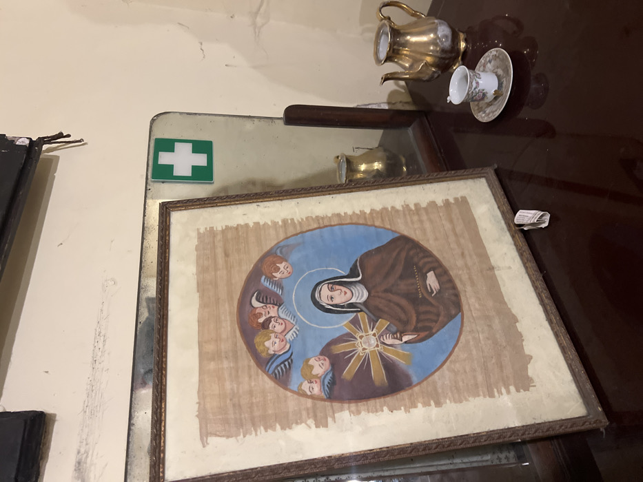 The image shows a framed painting depicting a woman wearing a brown robe, holding a staff with a glowing orb in her hand, surrounded by angels. The painting is hanging on a wall in a room with a mirror and a wooden dresser. On the dresser, there is a teapot and a small teacup. The room is located in Napoli, Italy, at the coordinates 40.852 latitude and 14.257 longitude. The image depicts the interior of what may be a home or a religious space. There is a green sign with a white plus symbol attached to the wall. There is a paper tag visible at the bottom of the frame of the painting.
