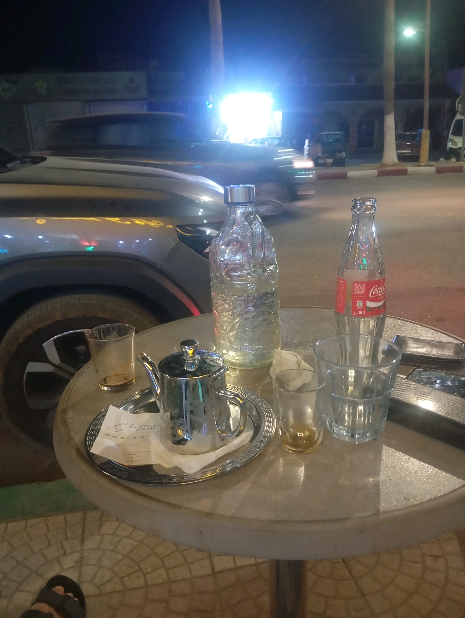 The photograph depicts an outdoor cafe scene at night, likely in Boufakrane, Morocco, as indicated by the presence of a Coca-Cola bottle with Arabic text. In the foreground, a round, light-colored table holds several items. To the left, a small, clear glass contains a amber-colored liquid. Next to it, a silver teapot with a lid sits on a silver tray with an ornate rim. A folded white paper napkin rests on the tray. To the right of the teapot, a glass bottle of Coca-Cola, with its iconic red label, is standing. Behind the Coca-Cola bottle, another clear glass, this one larger and with ridged detailing, stands empty. Partially visible to the right of the Coca-Cola bottle is a metallic object, possibly an ashtray.
In the middle ground, the front of a silver car is prominently displayed. The car appears to be parked, with its headlights reflecting on the wet or shiny road surface. The car's wheel is visible, showing a complex rim design.
The background is a dimly lit street scene. Buildings with arched doorways and windows are visible, some illuminated by artificial light. A tall, slender pole, likely a streetlight, with a bright light at its top, dominates the right side of the background. Trees are also present. The overall atmosphere is that of a casual evening gathering at a roadside cafe, with the sounds and sights of urban life implied. A portion of a person's foot wearing a sandal is visible in the bottom left corner of the image, suggesting the photographer or a companion is present.