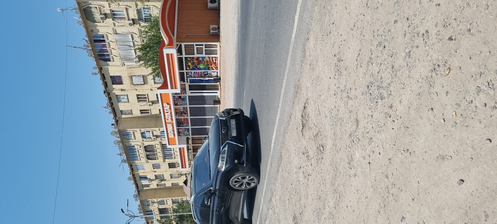 In Ismoili Somoni, Tajikistan, a black Lexus SUV is parked on the side of a paved road. The SUV has a license plate that reads "77000510". In the background, there is a light-colored, multi-story apartment building with numerous satellite dishes on its roof. In front of the apartment building, there is a small, modern-looking store with an orange awning and large glass windows displaying various items. The ground in the foreground is unpaved and covered with gravel and dirt. The sky is clear and blue.
