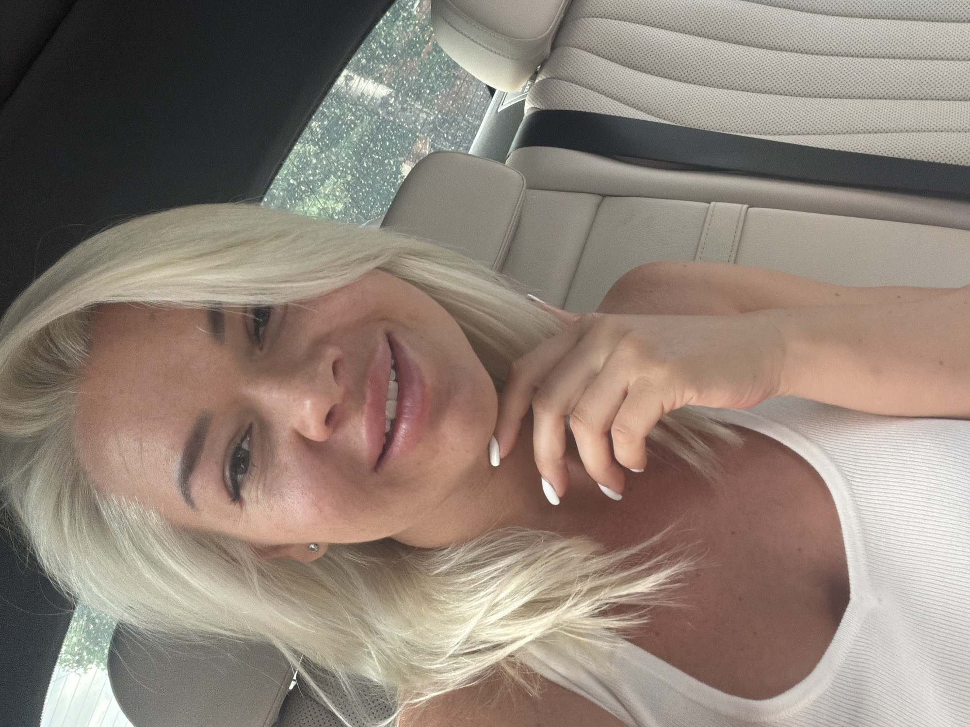 A woman with blonde hair is in the back seat of a car. She is wearing a white tank top and has white nail polish. The car has light colored leather seats. The city is Unknown, Unknown.