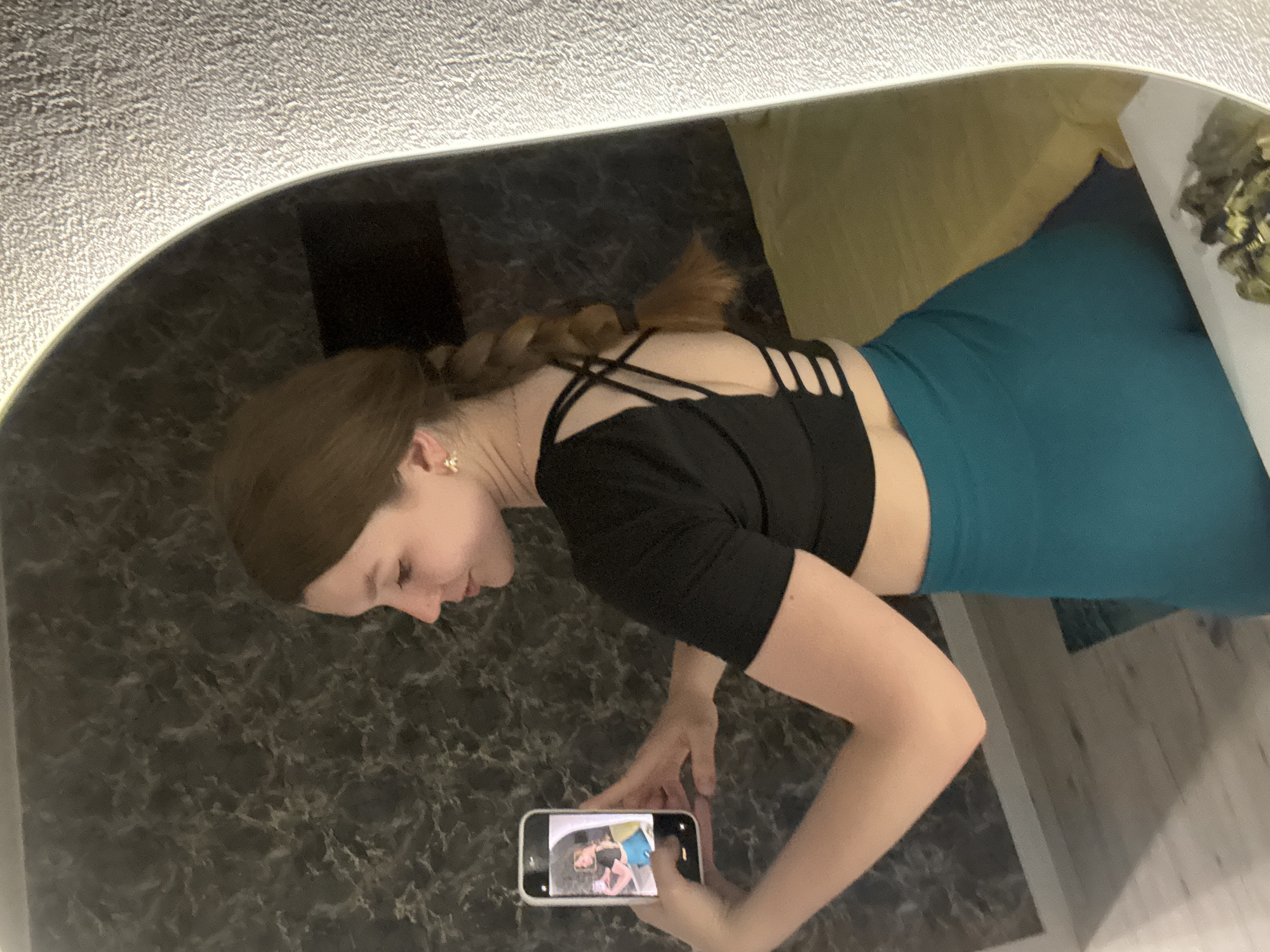 A woman is taking a picture of herself in a mirror with her smartphone. The mirror is large and has a rounded shape, and the woman is standing close to it. She is wearing a black top with a strappy back and teal leggings. Her hair is styled in a braid. The background includes a patterned wall and a portion of a room with a pillow and possible foliage. There is no visible information in the image to confirm its location in the city of Izhevsk, Russia.