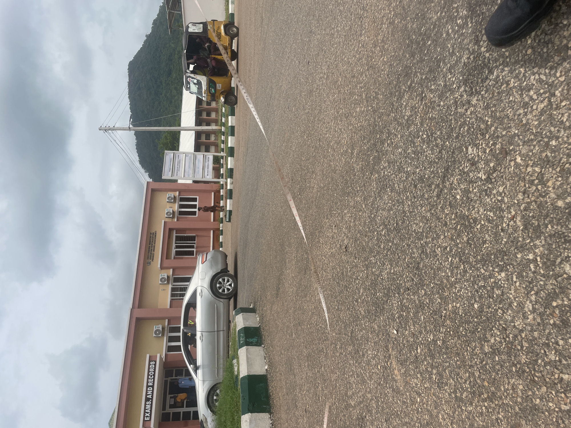 This media file captures a street scene in Lokoja, Nigeria. The perspective is from the ground level, looking upwards and to the side, suggesting the photographer might be standing or sitting. The foreground is dominated by a gray asphalt road with visible pebbles and some scattered debris. A white tape with red markings, possibly a measuring tape, stretches diagonally across the road.
On the left side of the frame, a light-colored sedan is parked alongside a building. The building, painted in shades of beige and pink, appears to be an administrative or institutional structure. It has multiple windows with dark frames and several air conditioning units mounted on its facade. A sign above the entrance reads "EXAMS. AND RECORDS." Another, larger sign is mounted on a pole in front of the building, with text indicating it's related to the "FEDERAL MINISTRY OF CATION REVITALIZATION PROJECT (NEEDS ASSESSMENT)" and "BA-HAS LIMITED."
Further down the road, a yellow three-wheeled vehicle, commonly known as a "Keke Napep" or "Marwa," is visible. It appears to be carrying passengers. In the background, a lush green, tree-covered mountain rises against a cloudy sky. The overall impression is of a bright, possibly humid day, typical of a tropical climate. The presence of the Keke Napep, the style of the building, and the mountain backdrop are strong indicators of a Nigerian location. There are a few figures visible, including a person near the car and others in or around the yellow vehicle, but their details are not clear. The scene appears to be of daily life and infrastructure in Lokoja.