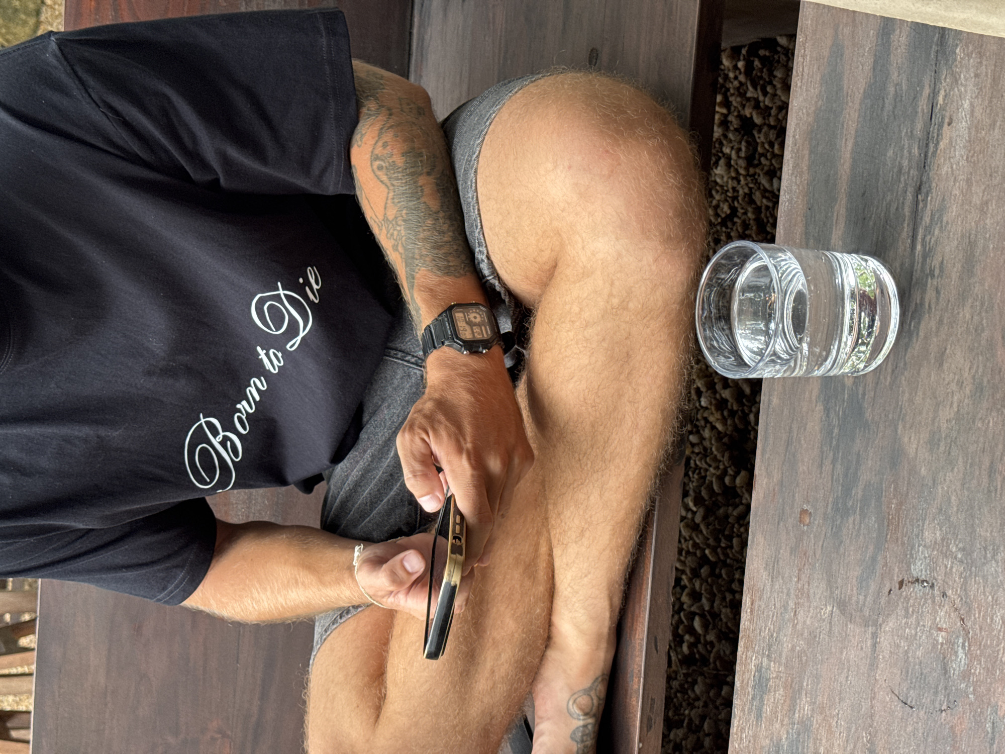 In Ahangama, Sri Lanka, a person is seated cross-legged on a wooden bench. They are wearing a black t-shirt with white text that reads "Born to Die" and grey shorts. Their arms are crossed, and they are holding a smartphone in their right hand, looking at the screen. A watch with a black band and digital display is visible on their left wrist. A glass of clear liquid, likely water, sits on a wooden surface in front of them. The background shows a blurred natural environment with rocks and foliage.