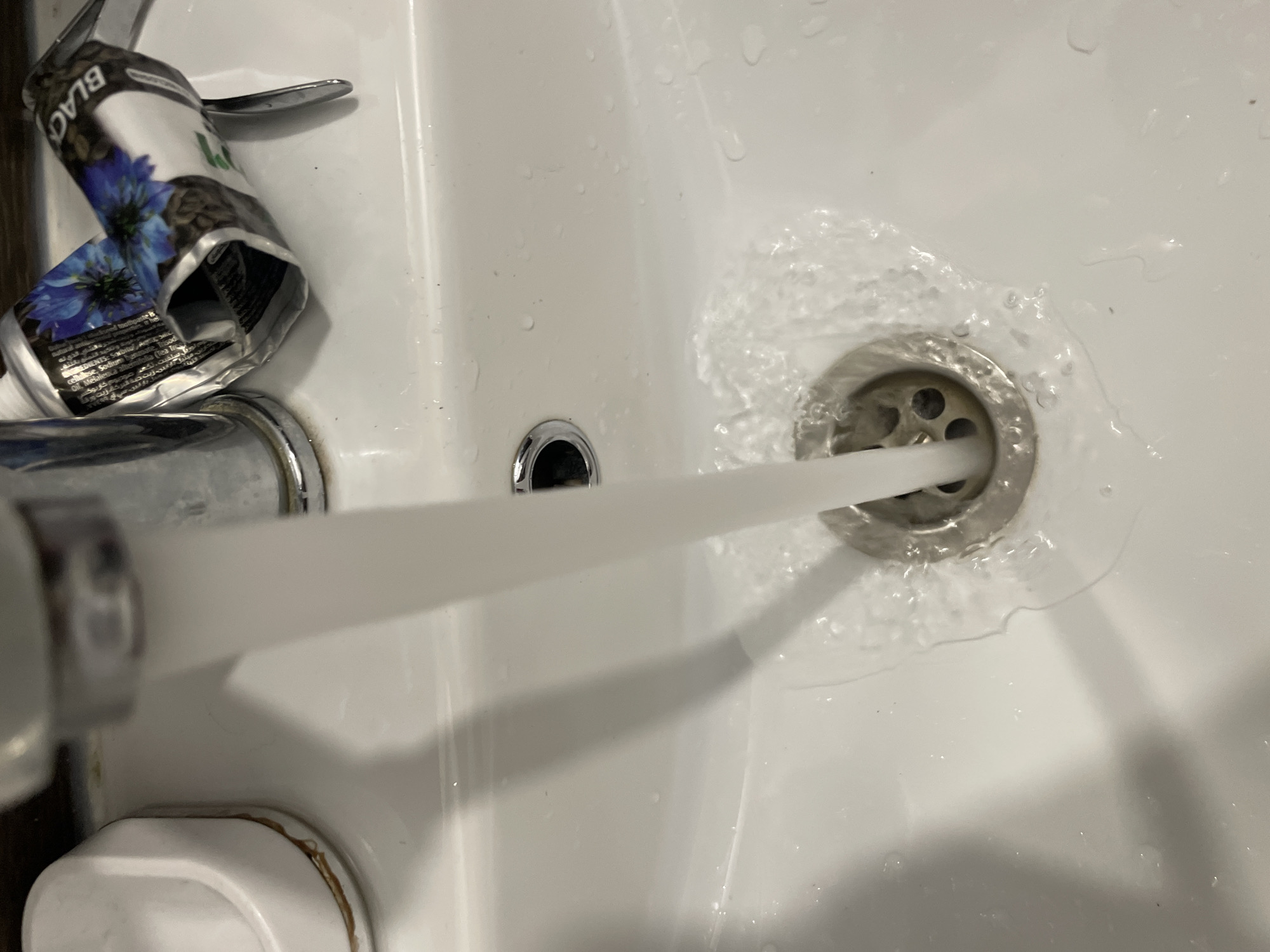 A close-up, overhead view shows a stream of water flowing from a faucet into a white ceramic sink. The water is hitting a chrome drain in the center of the sink, creating ripples and splashing effects. To the left of the faucet, a white soap dispenser with a brown stain around its base is visible. To the right of the faucet, a crumpled tube of what appears to be a cosmetic product with blue flowers and the word "BLACK" printed on it rests on the sink's edge. The surrounding sink surface is wet, with several small water droplets scattered across it. The image does not provide any specific details related to the city of Sinyavino, Russia.