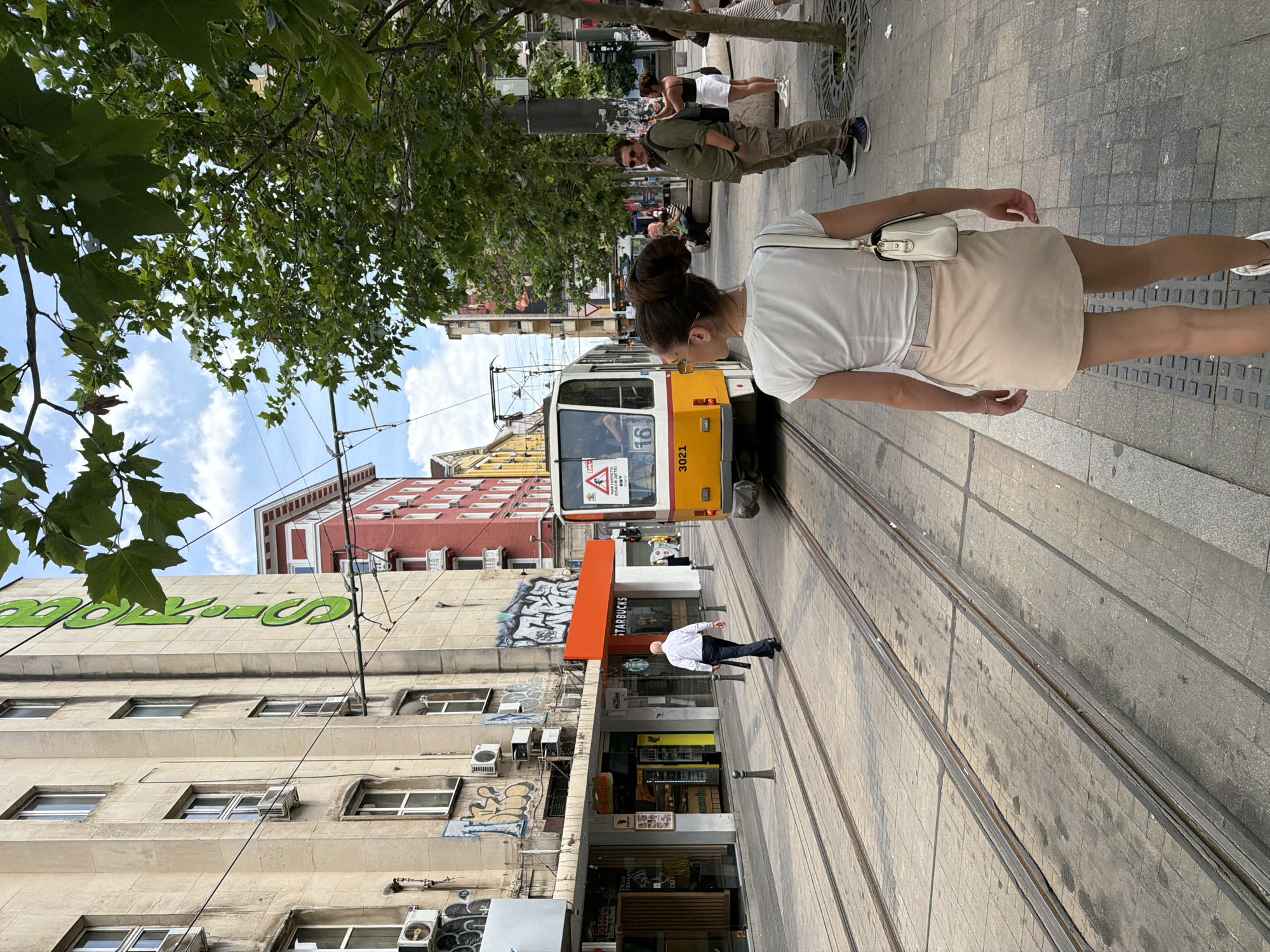The image depicts a street scene in Sofia, Bulgaria, characterized by tram tracks running down the center of the road. A yellow and white tram is visible, heading away from the viewer, with the number 3021 displayed. To the left of the tram tracks, a man in a white shirt and dark trousers walks away from the camera. Along the left side of the street, a light-colored building with multiple windows and air conditioning units is visible. Graffiti adorns the lower facade of this building, and an orange awning extends over a storefront. Further down the street, a red building with white balconies stands. On the right side of the street, a woman with her hair in a bun, wearing a white t-shirt and a light-colored skirt, is seen from behind, walking away from the tram. A man in a green shirt and cargo pants stands on the sidewalk, looking towards the tram. Several other people are present in the background, and trees line the right side of the sidewalk. The sky is partly cloudy.